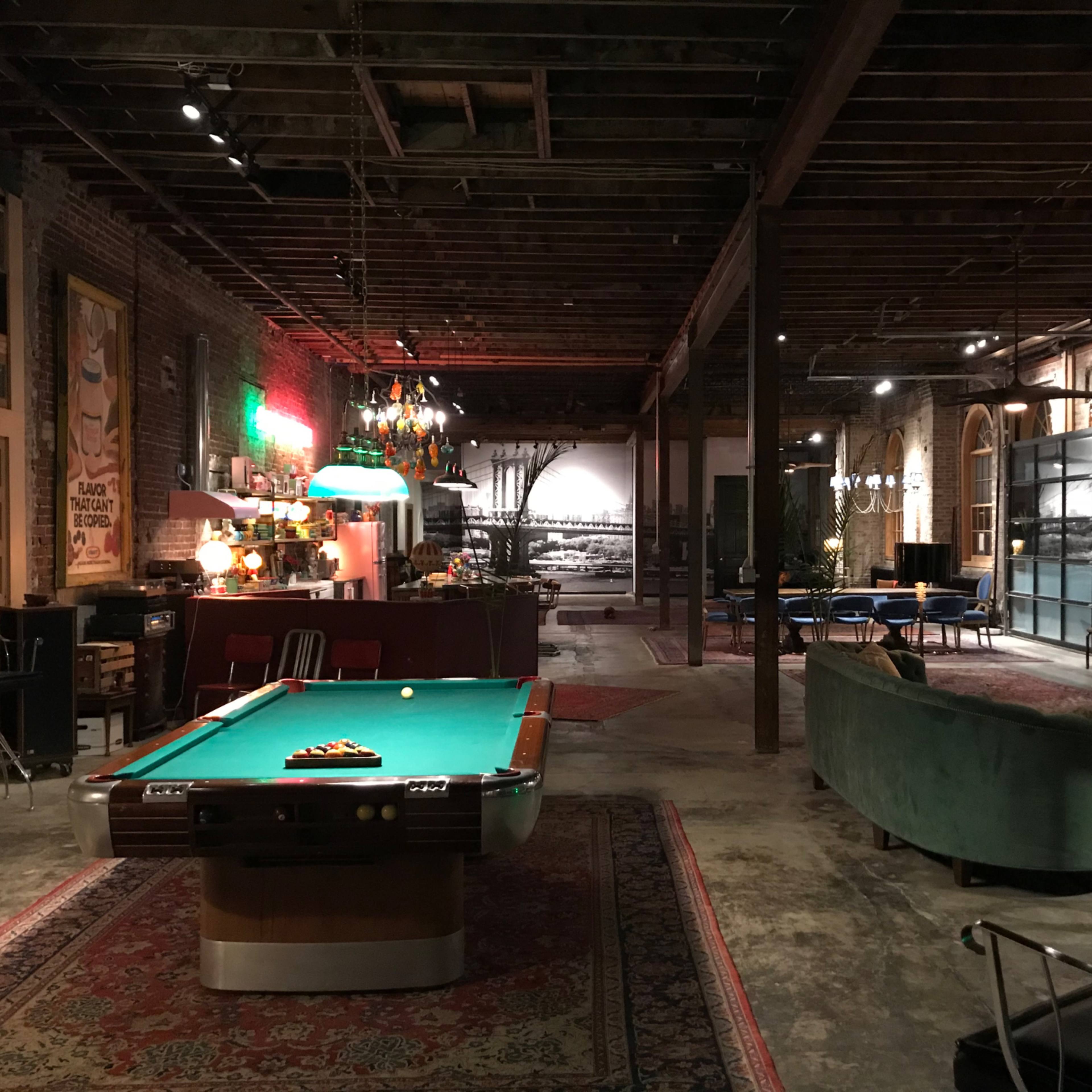 A spacious, industrial-style interior featuring a billiard table, vintage furniture, and exposed brick walls, illuminated by various light fixtures.