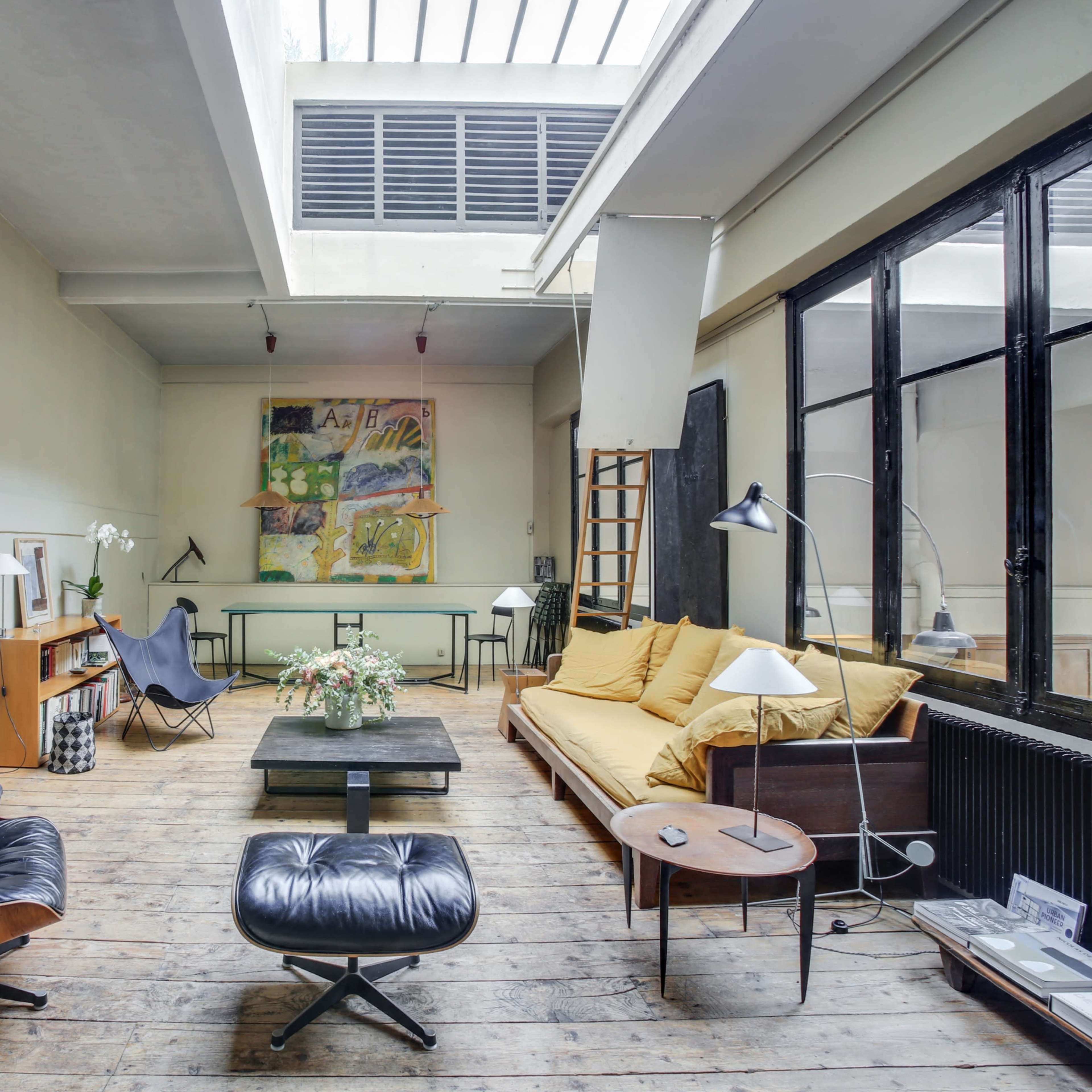 A spacious, modern living room with wooden flooring, large windows, a yellow sofa, and various pieces of furniture including an armchair and a coffee table.