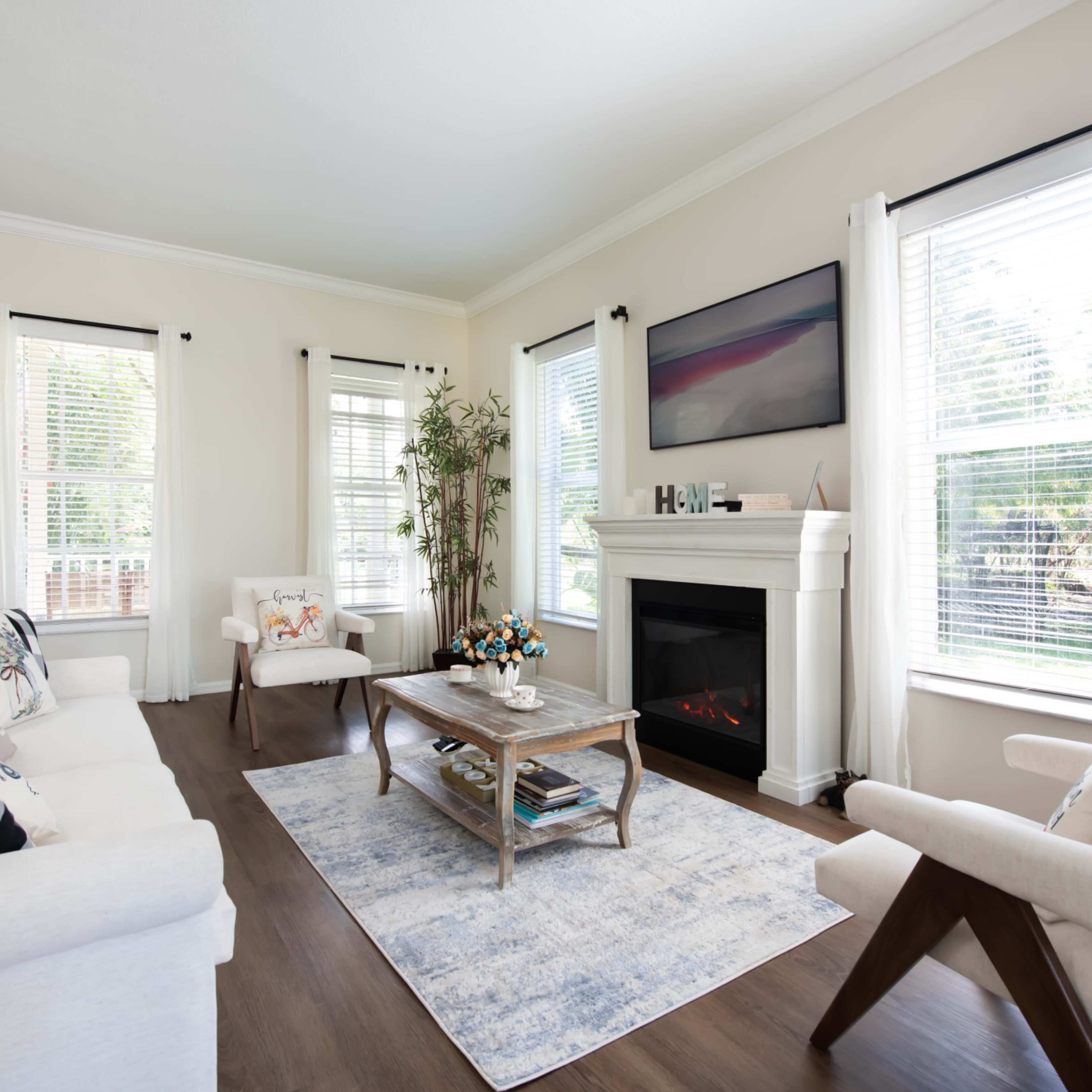 A bright living room features a white sofa and armchair, a central coffee table, and a fireplace below a wall-mounted television, with large windows allowing natural light.