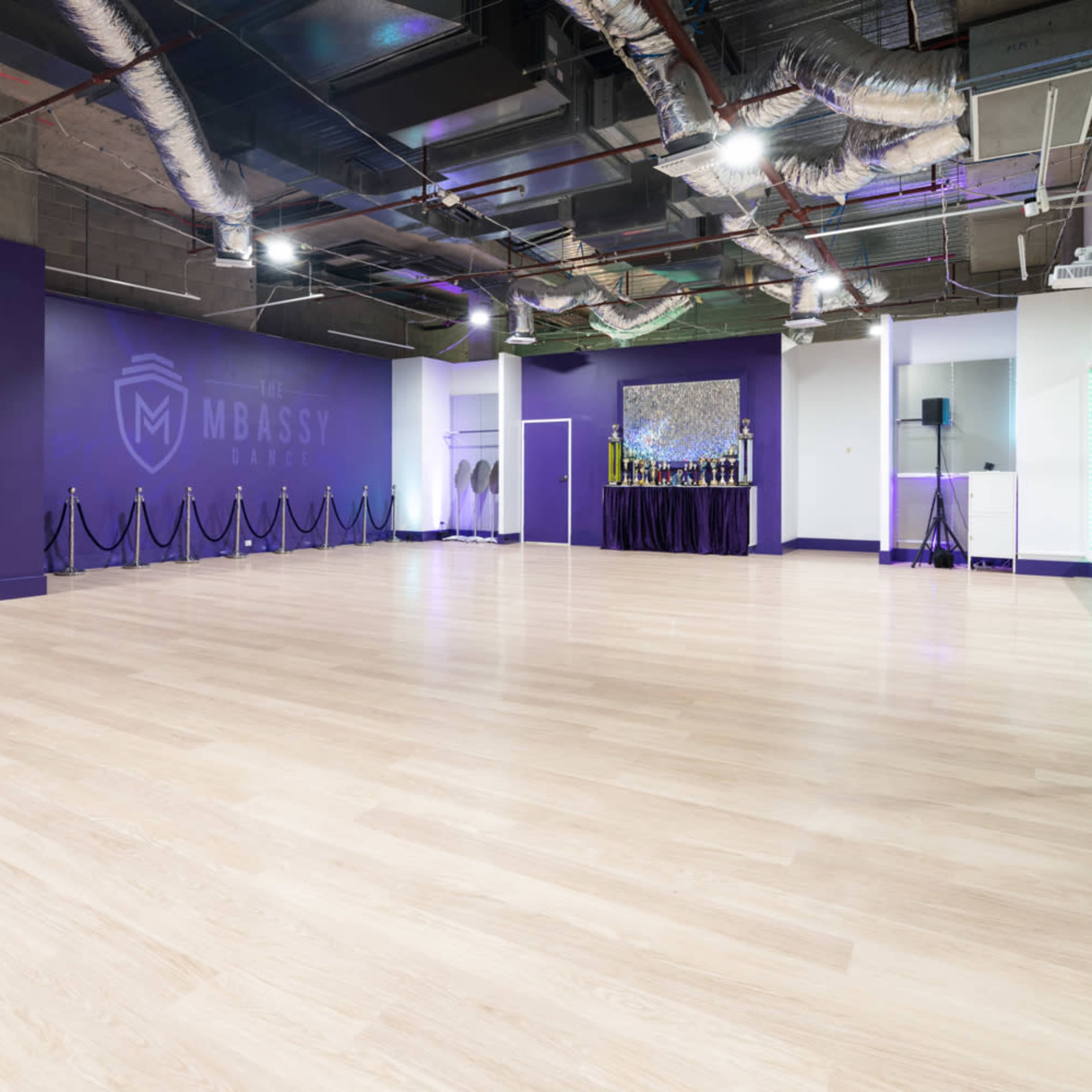 A spacious dance studio with a light wooden floor, purple walls, and mirrored panels.