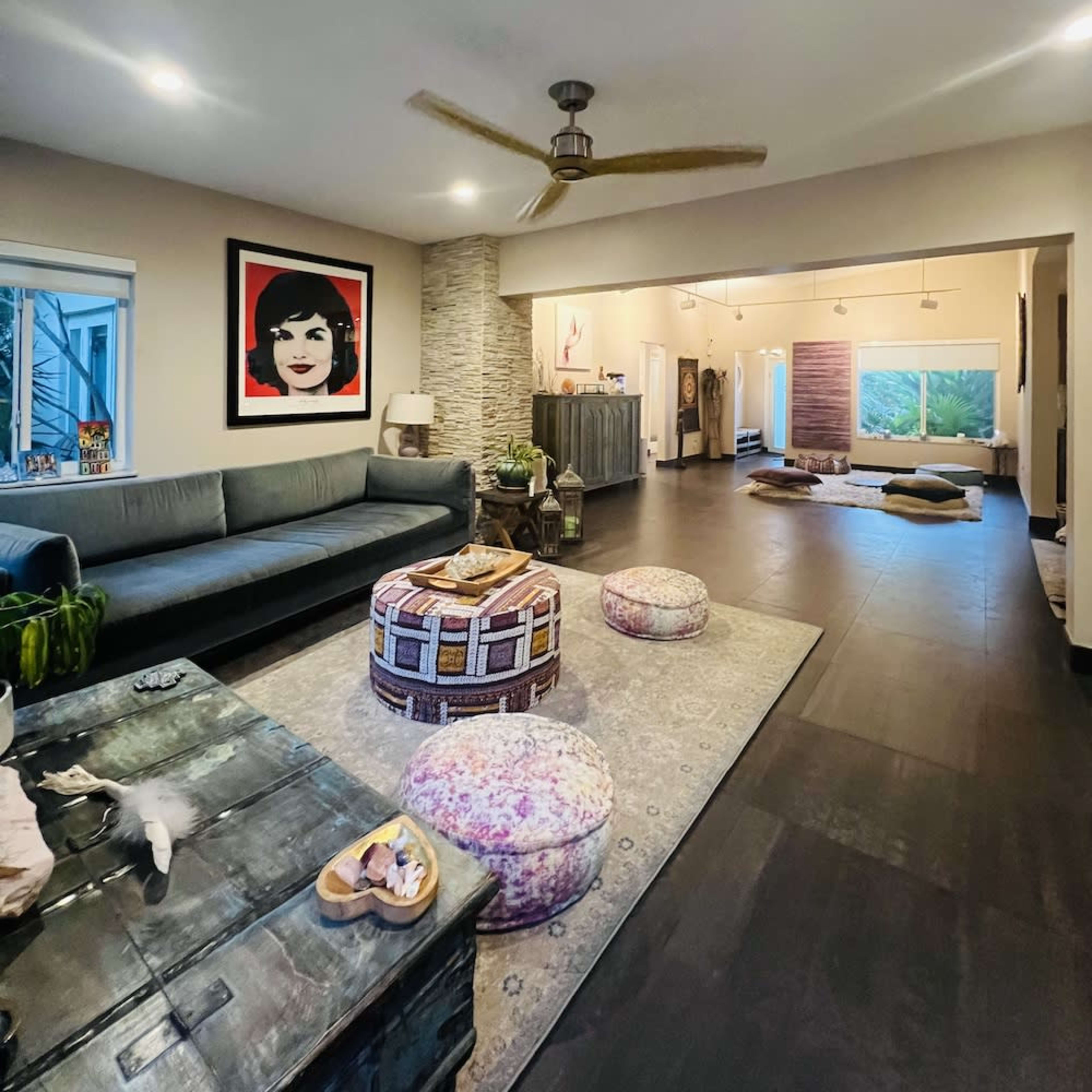 A spacious living room with a large sofa, a decorative rug, and a prominent artwork on the wall, leading into a well-lit area with additional seating and windows.