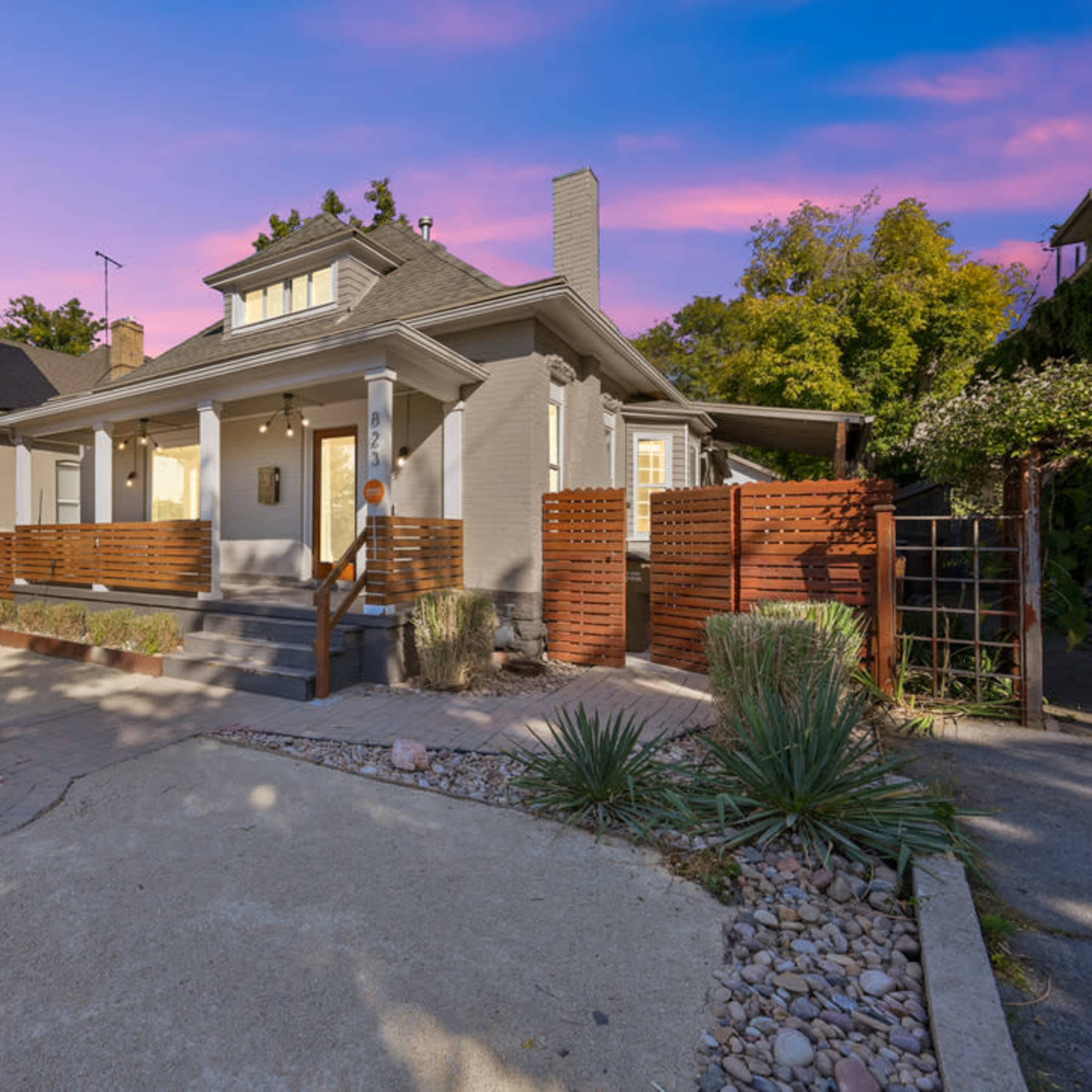 A gray single-story house with a front porch, surrounded by a landscaped yard and wooden fencing, set against a colorful sky at dusk.