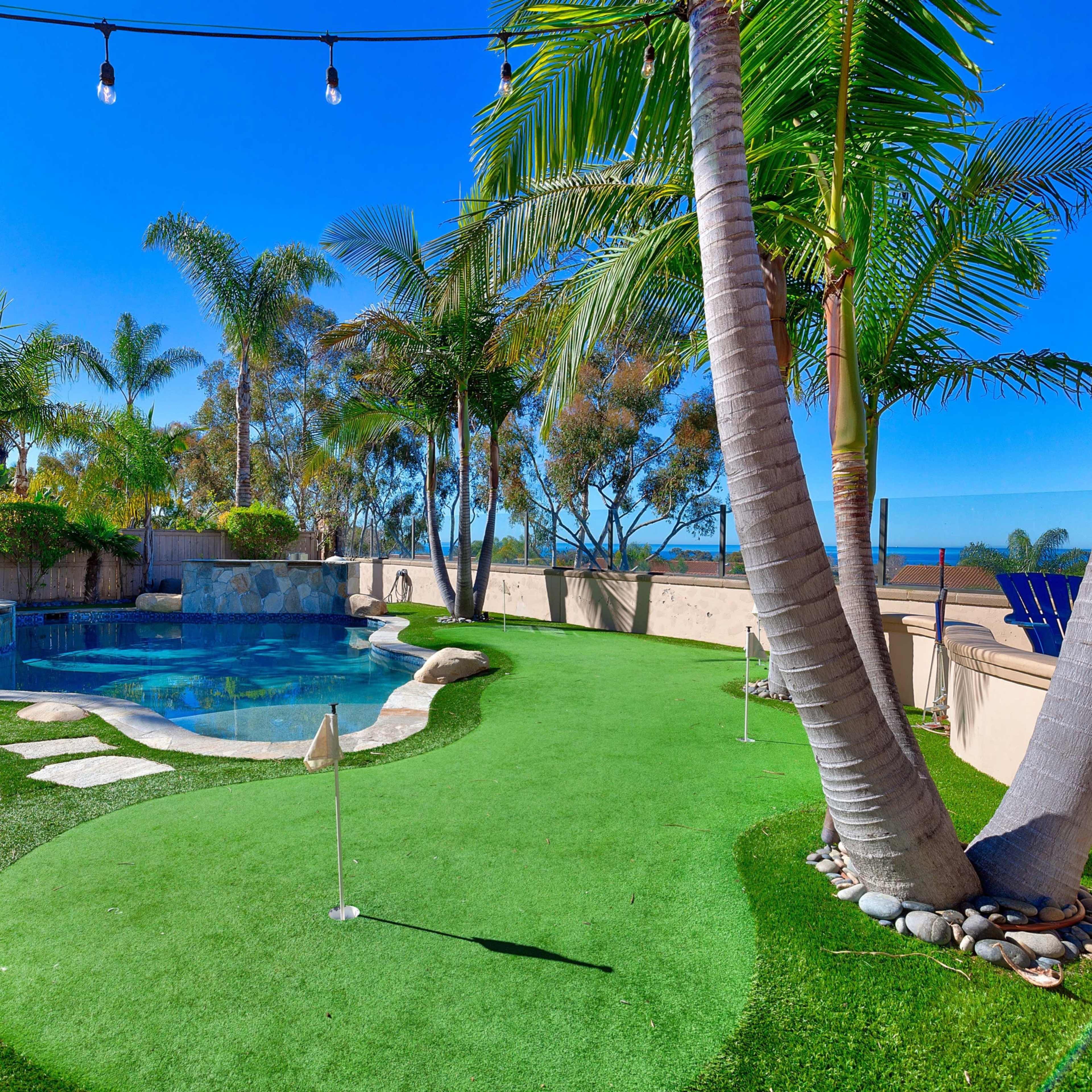 A landscaped backyard features a swimming pool surrounded by palm trees, artificial grass, and a small putting green.