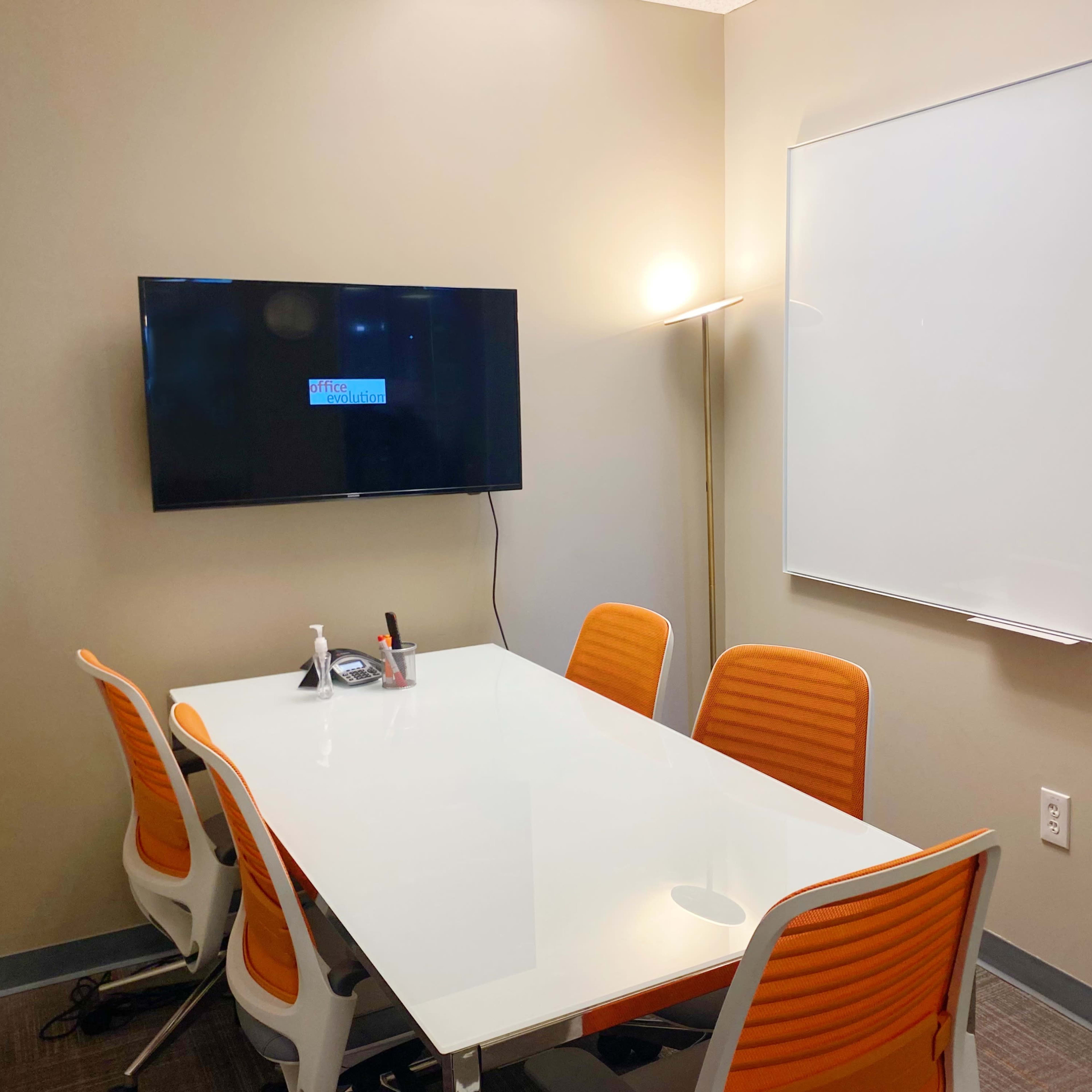A conference room features a glass table surrounded by orange chairs, a wall-mounted television, and a whiteboard.