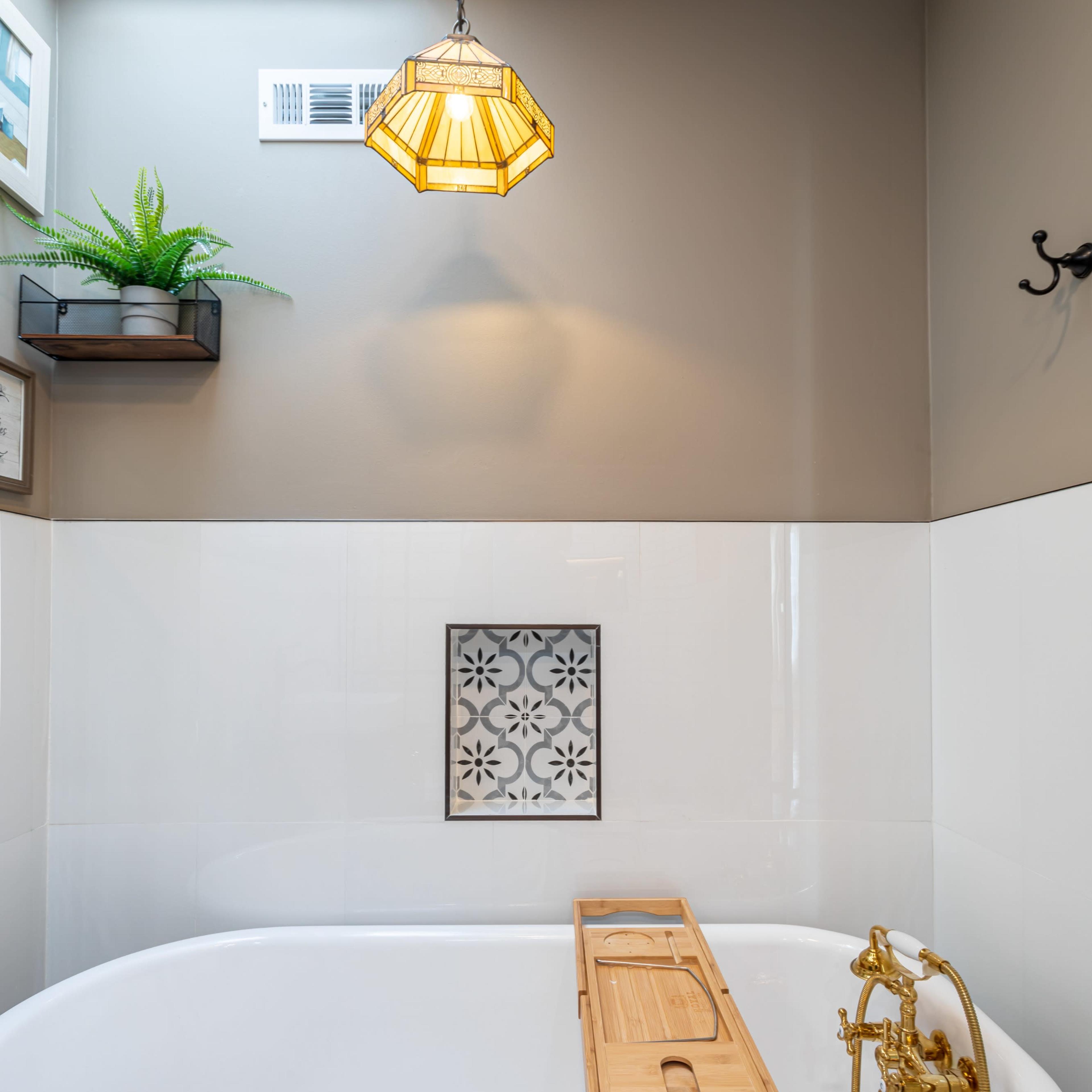 A vintage-style bathtub with a wooden tray on top is set against a wall featuring a framed decorative panel and a skylight above.