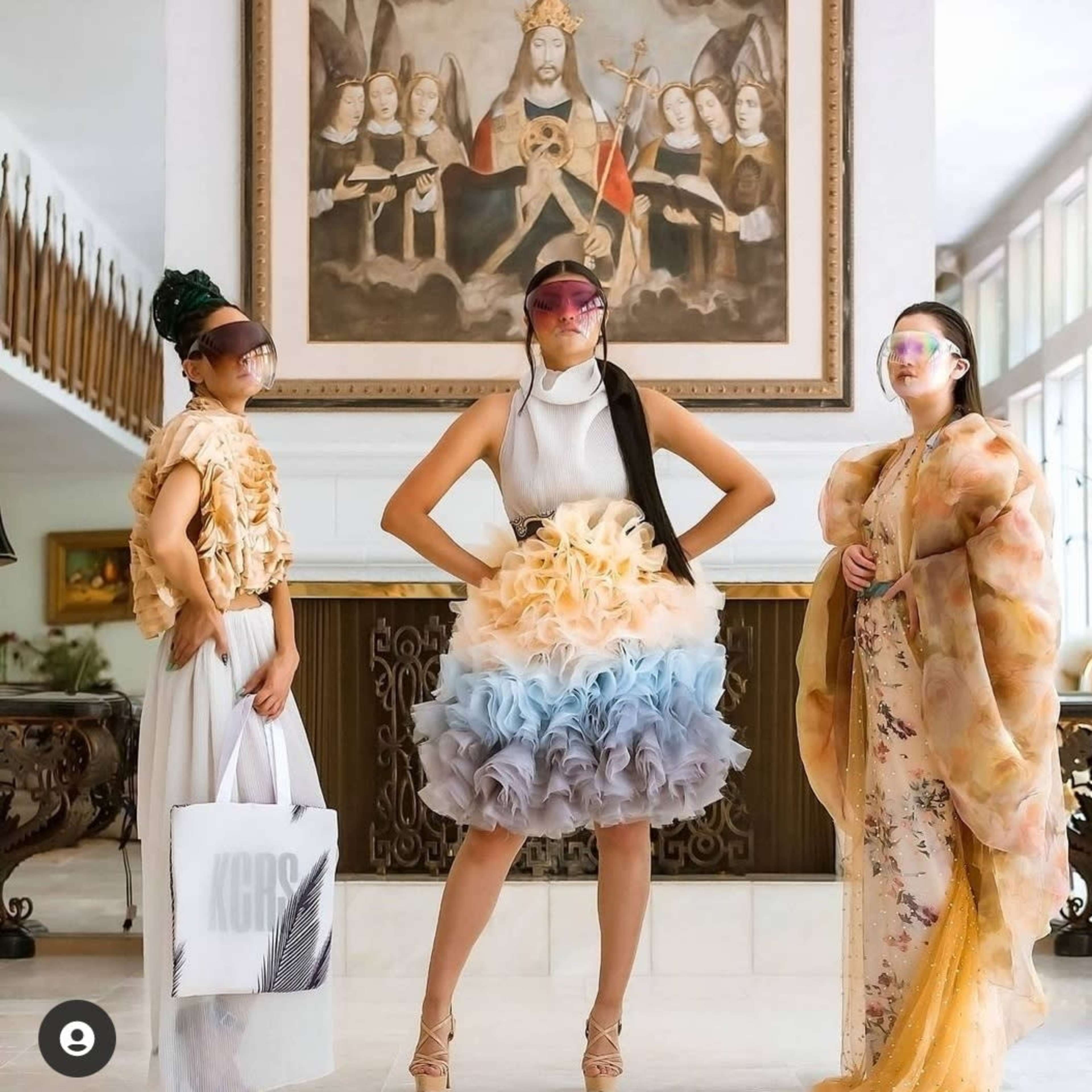 Three models pose in a spacious room with a large painting on the wall, each wearing unique fashion outfits and visors.