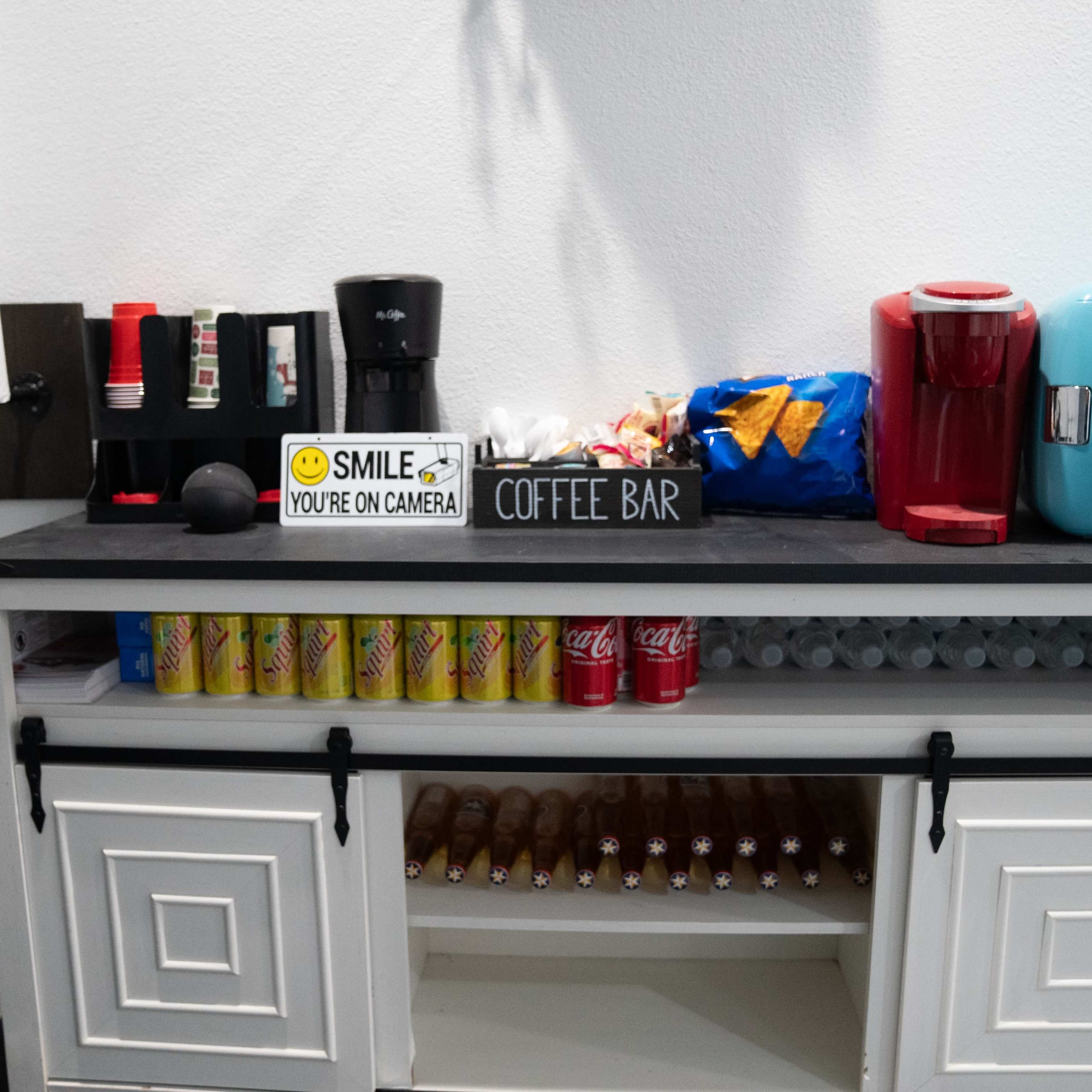 A coffee bar setup with a coffee maker, a snack assortment, and a cooler, accompanied by cans of soda and snacks neatly arranged on a black countertop.