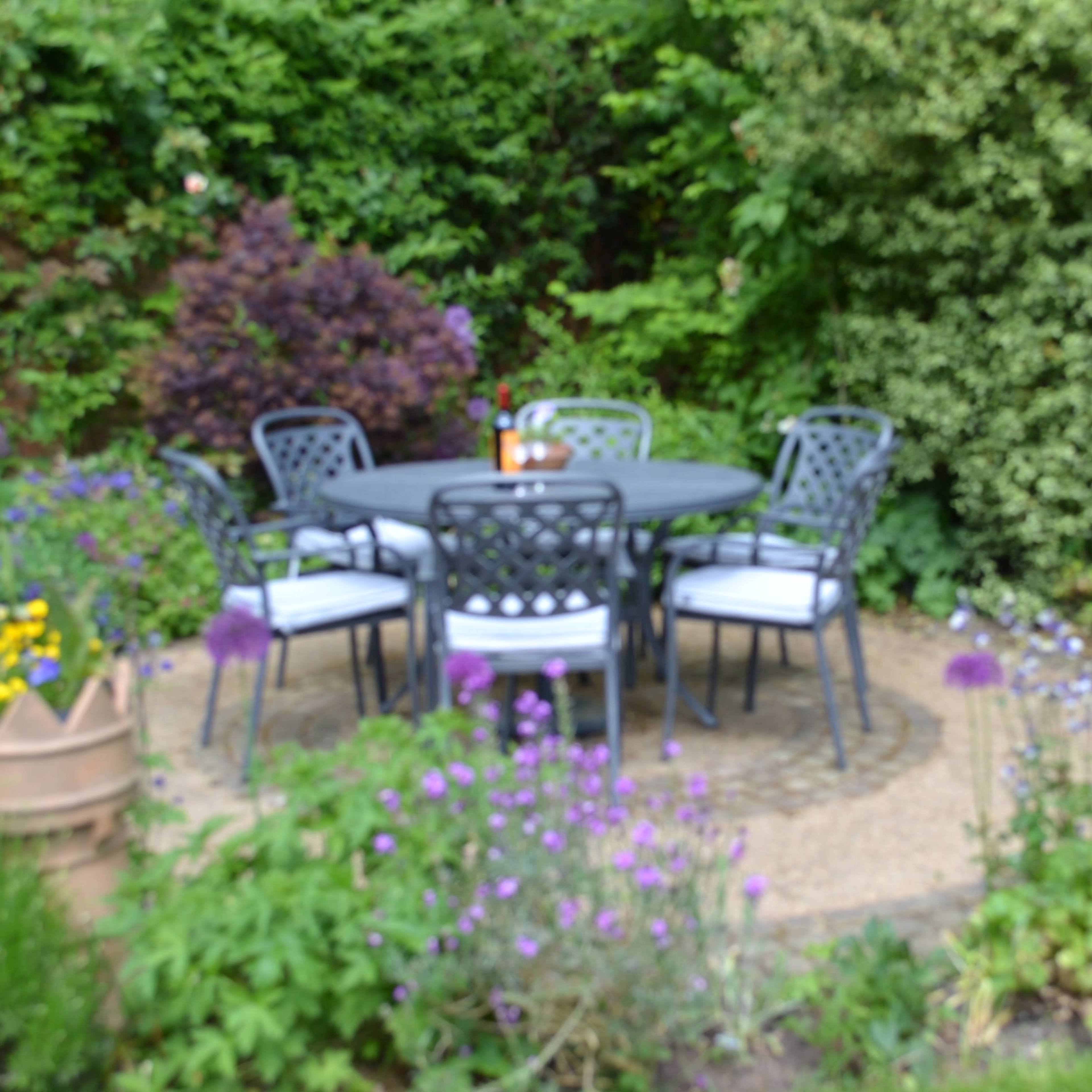 A circular table surrounded by four chairs sits on a stone path in a garden filled with various plants and flowers.