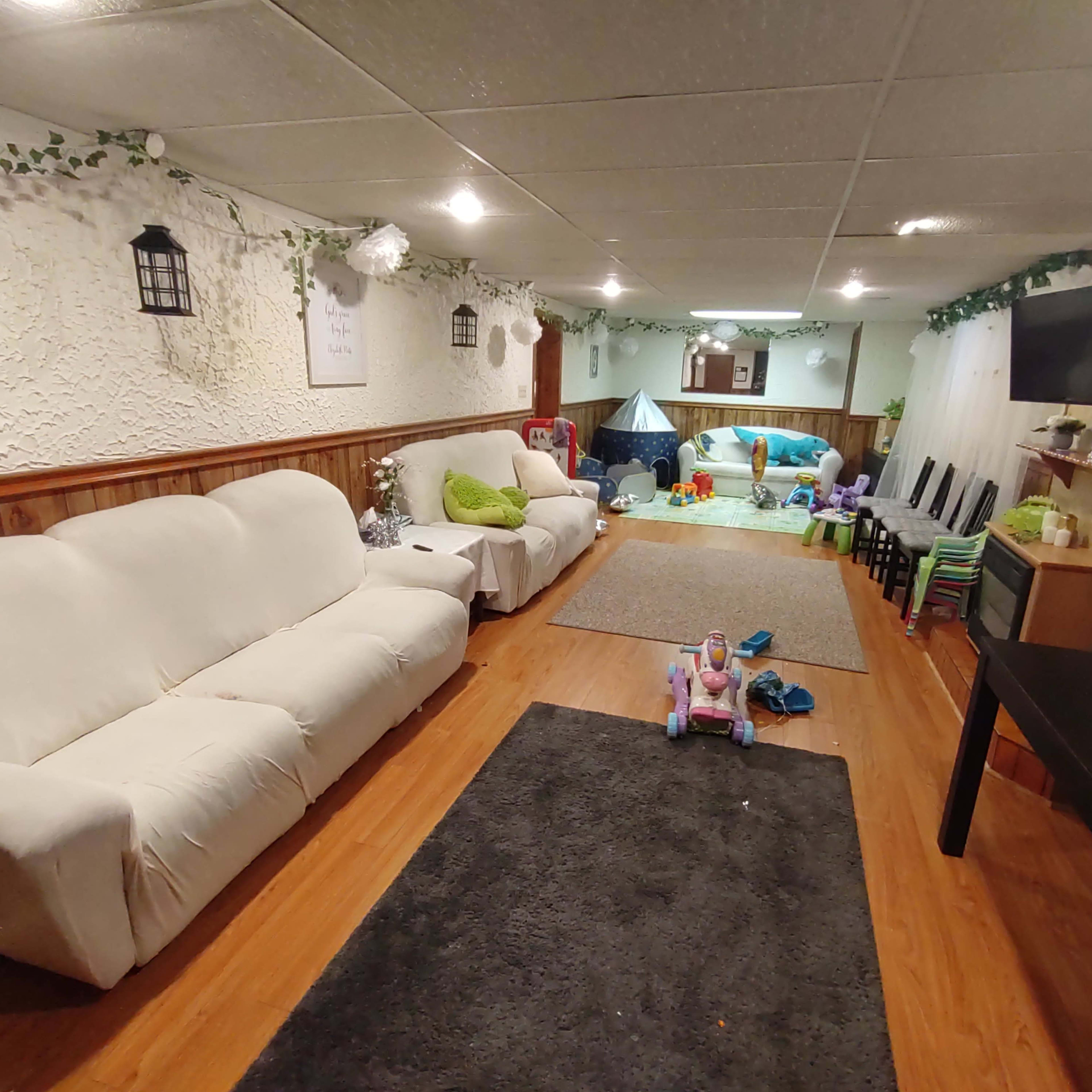 A spacious basement area furnished with white sofas, a black table, and various children's toys scattered across the floor.