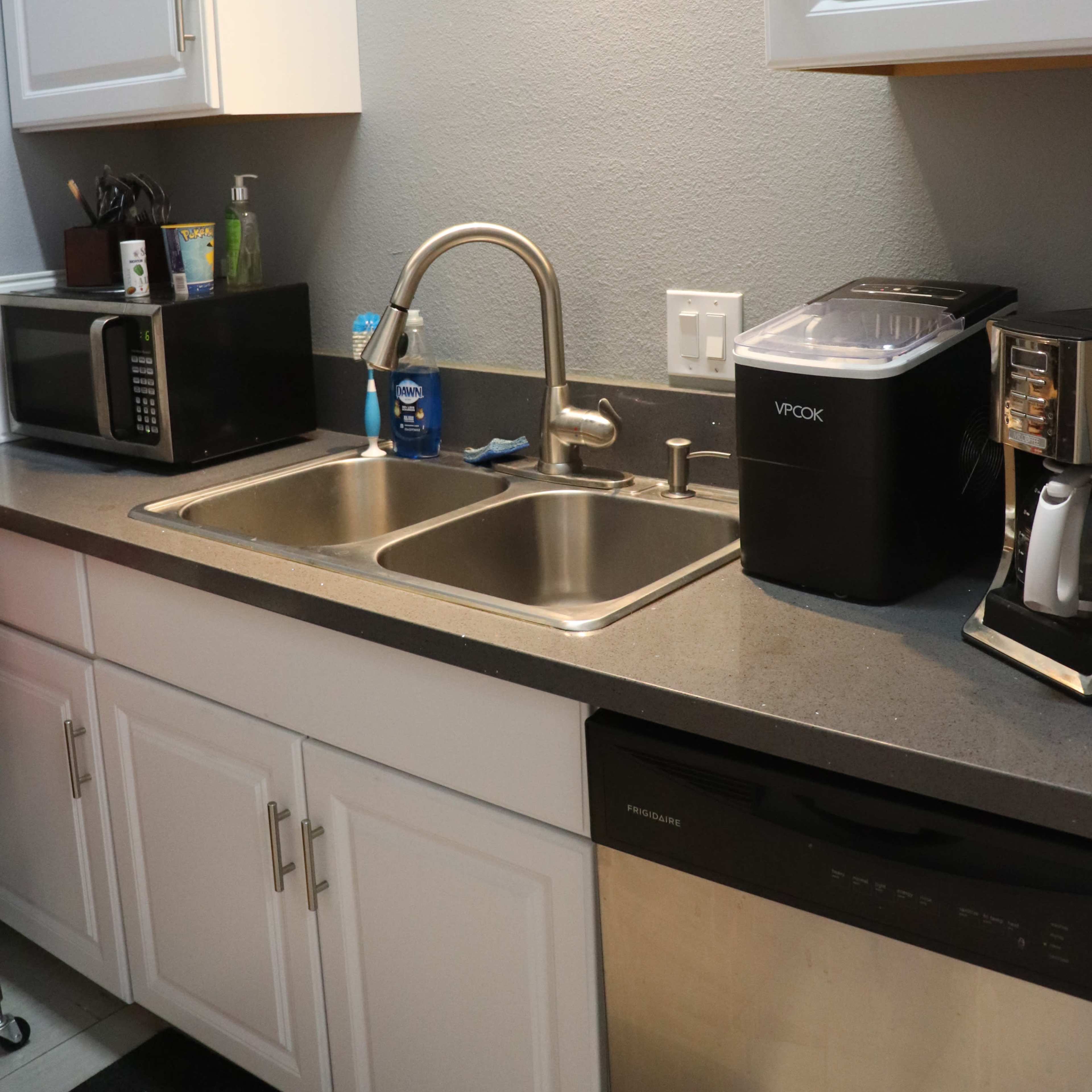 A kitchen sink area with two stainless steel sinks, a microwave, a countertop appliance, and a dishwasher beneath white cabinets.