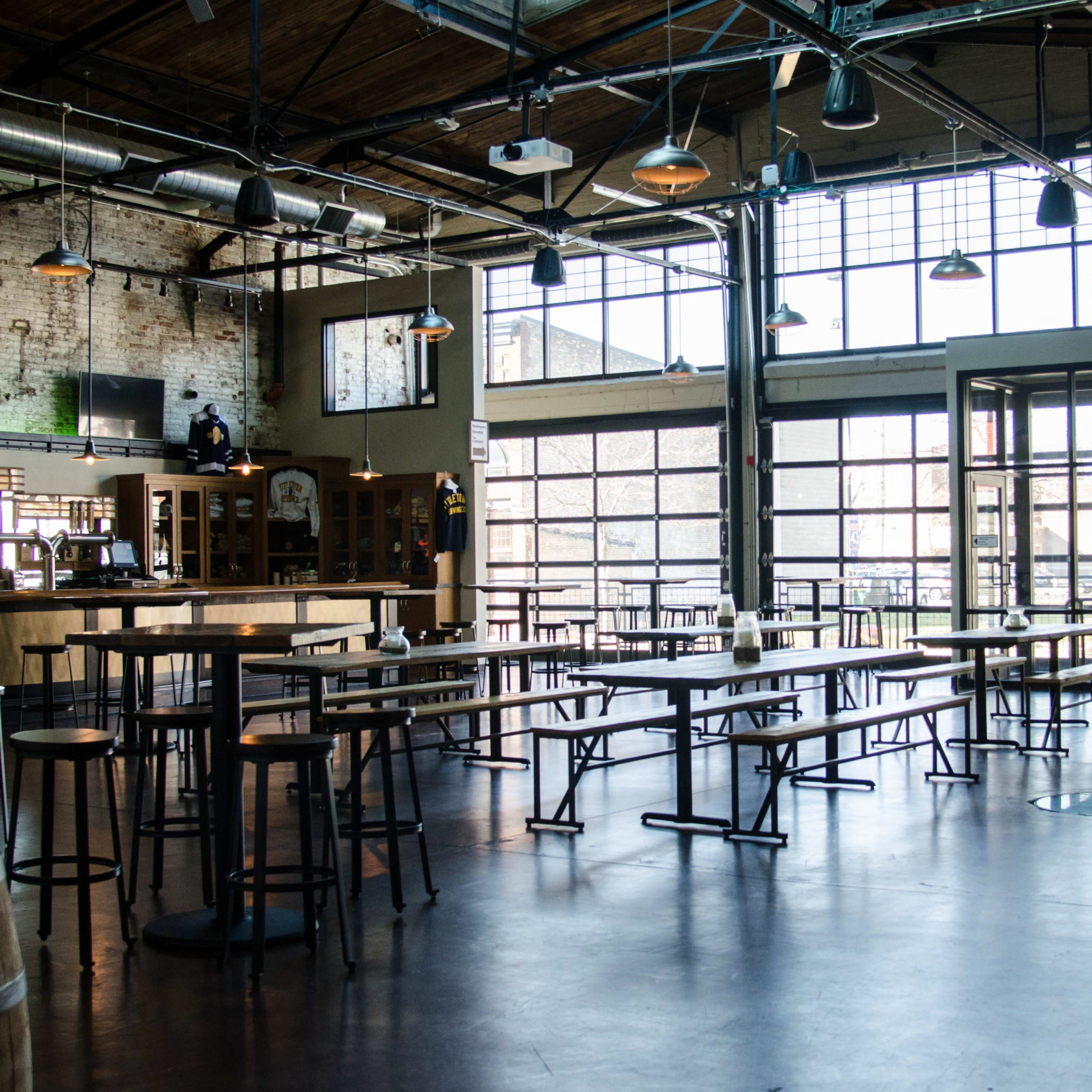 An industrial-style brewery interior with wooden tables, high stools, large windows, and a bar area.