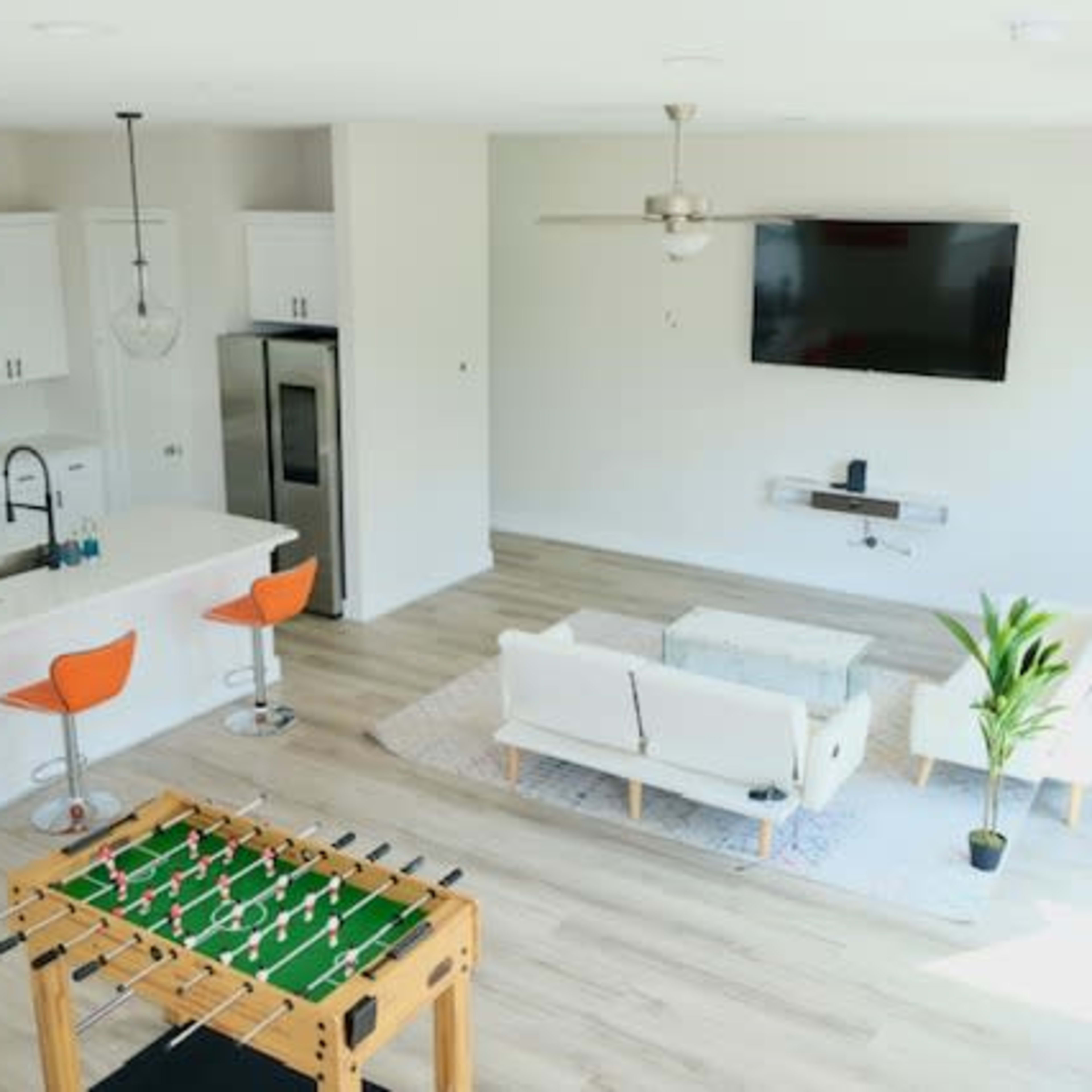 A modern living space showcases a kitchen with bar stools, a foosball table, a sofa set, and a wall-mounted television.
