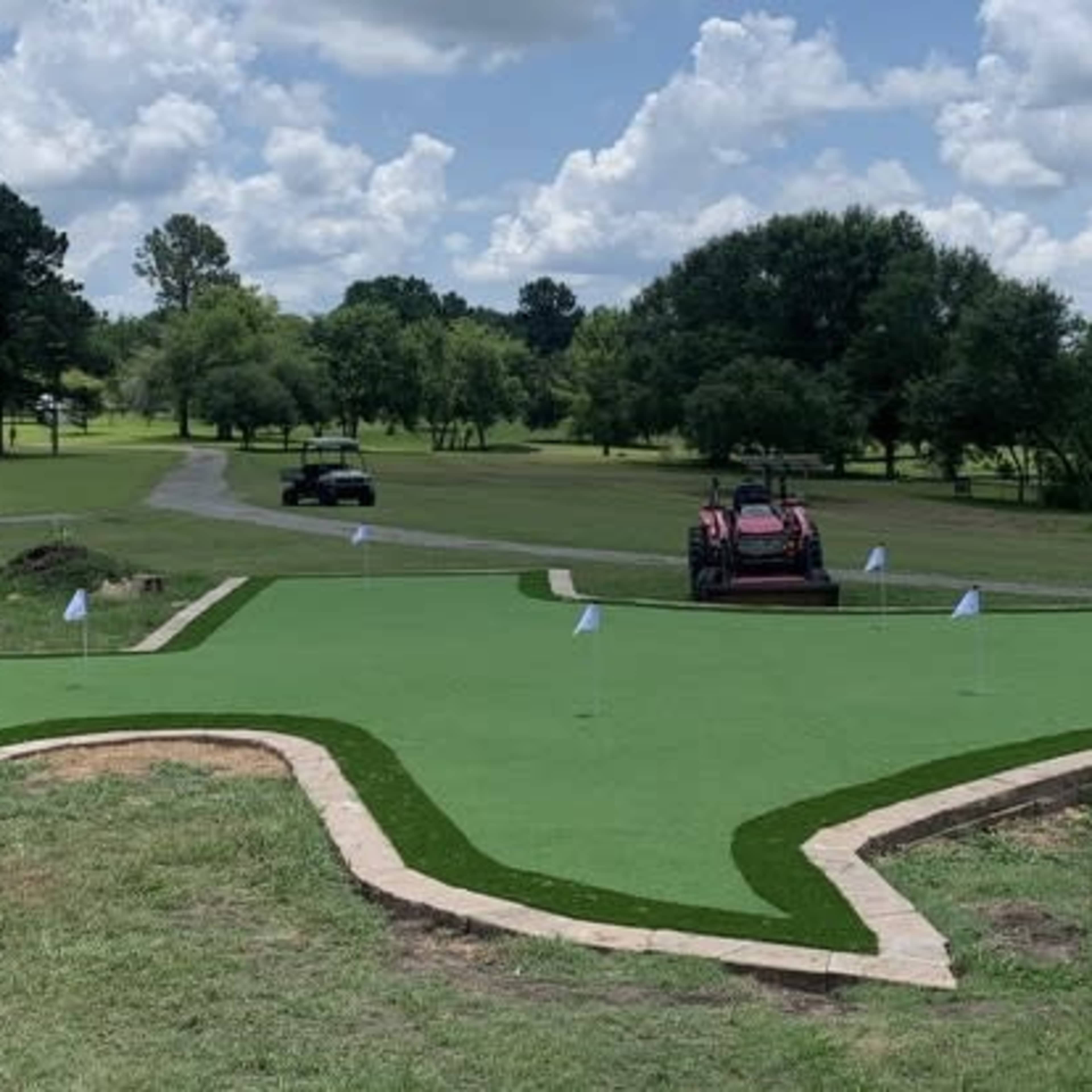 A Texas-shaped putting green surrounded by landscaping features, with a tractor parked nearby and a dirt mound at the edge of the green.