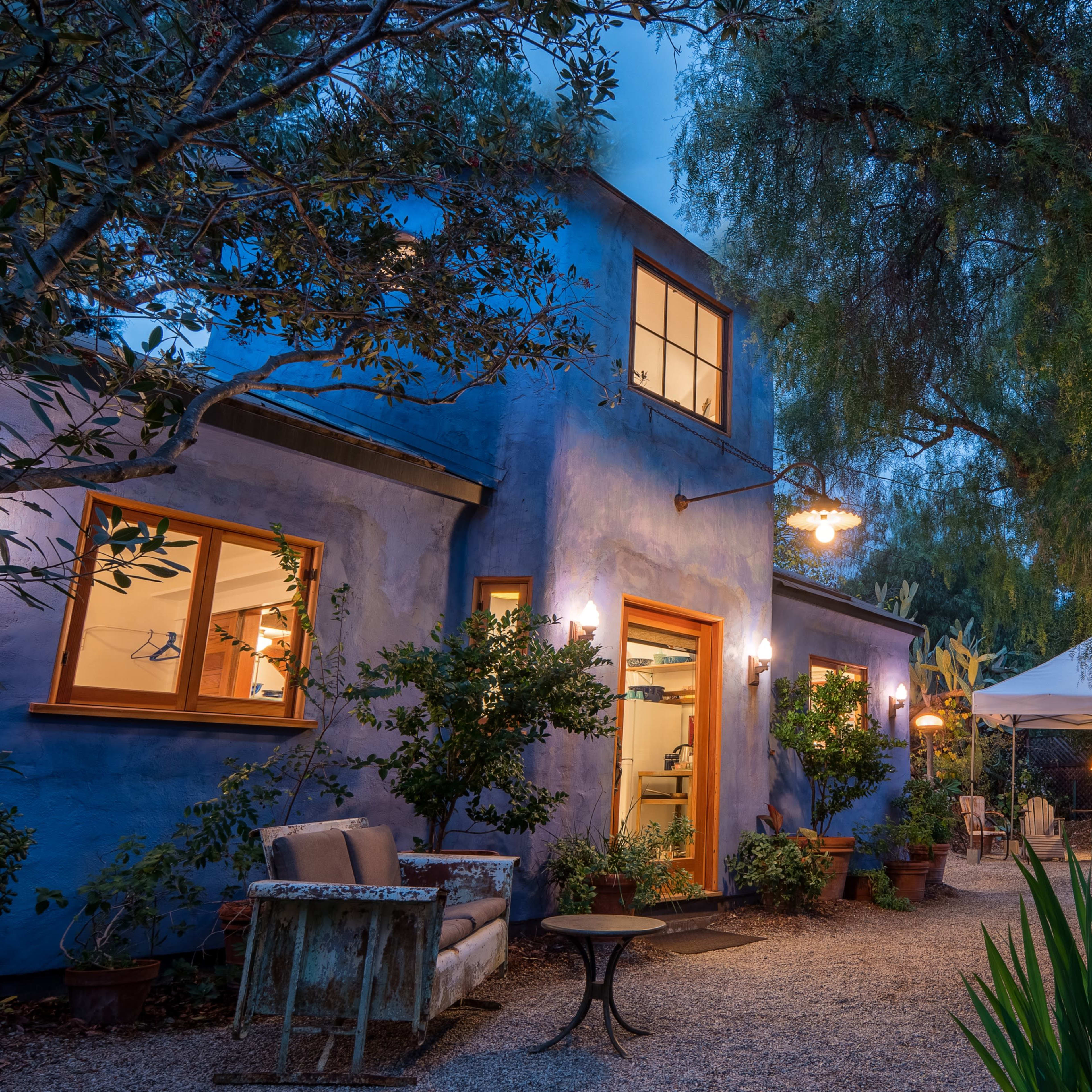 A blue-stucco house with illuminated windows is surrounded by greenery and outdoor seating under a canopy at dusk.
