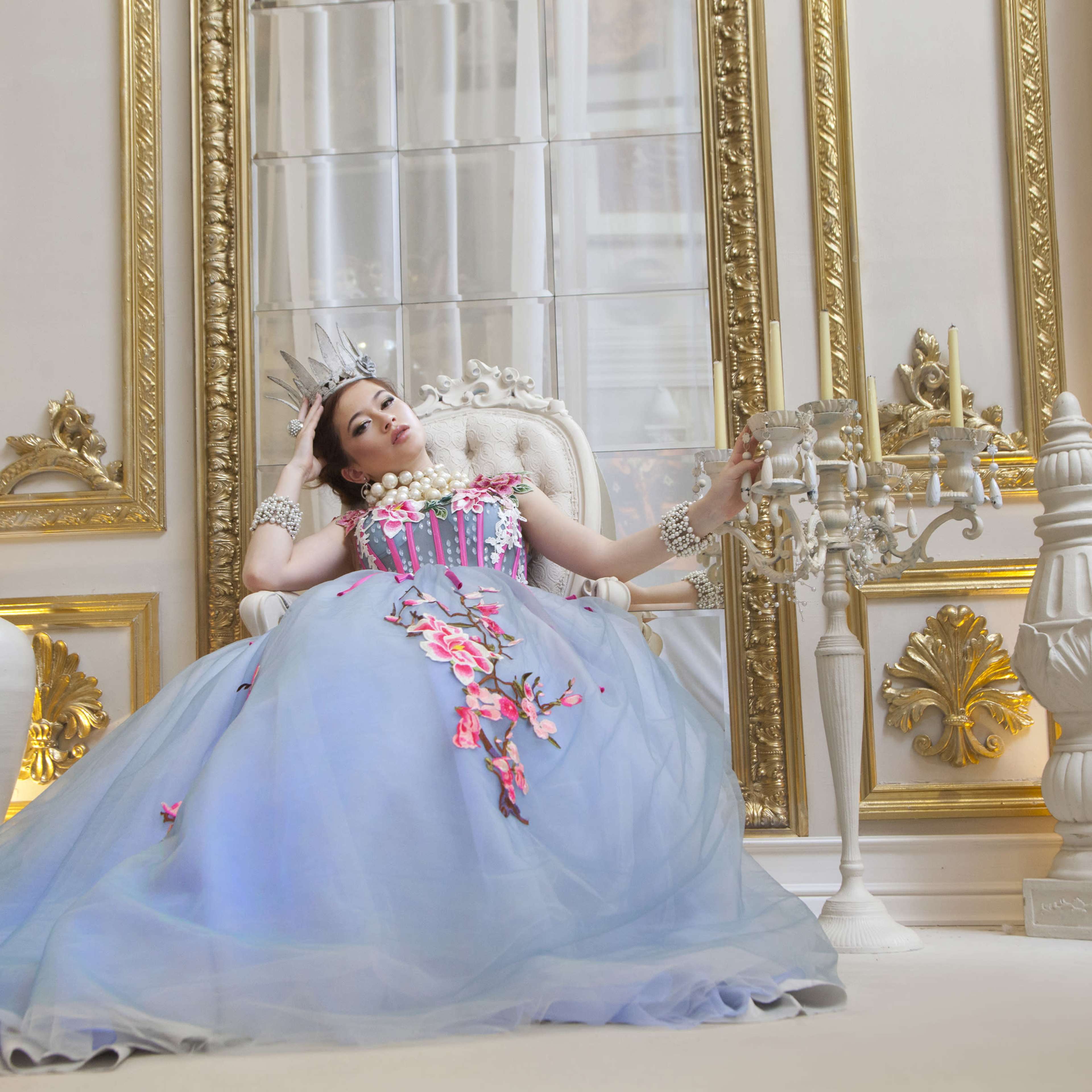 A woman wearing a blue gown adorned with floral decorations reclines elegantly in a richly decorated room.