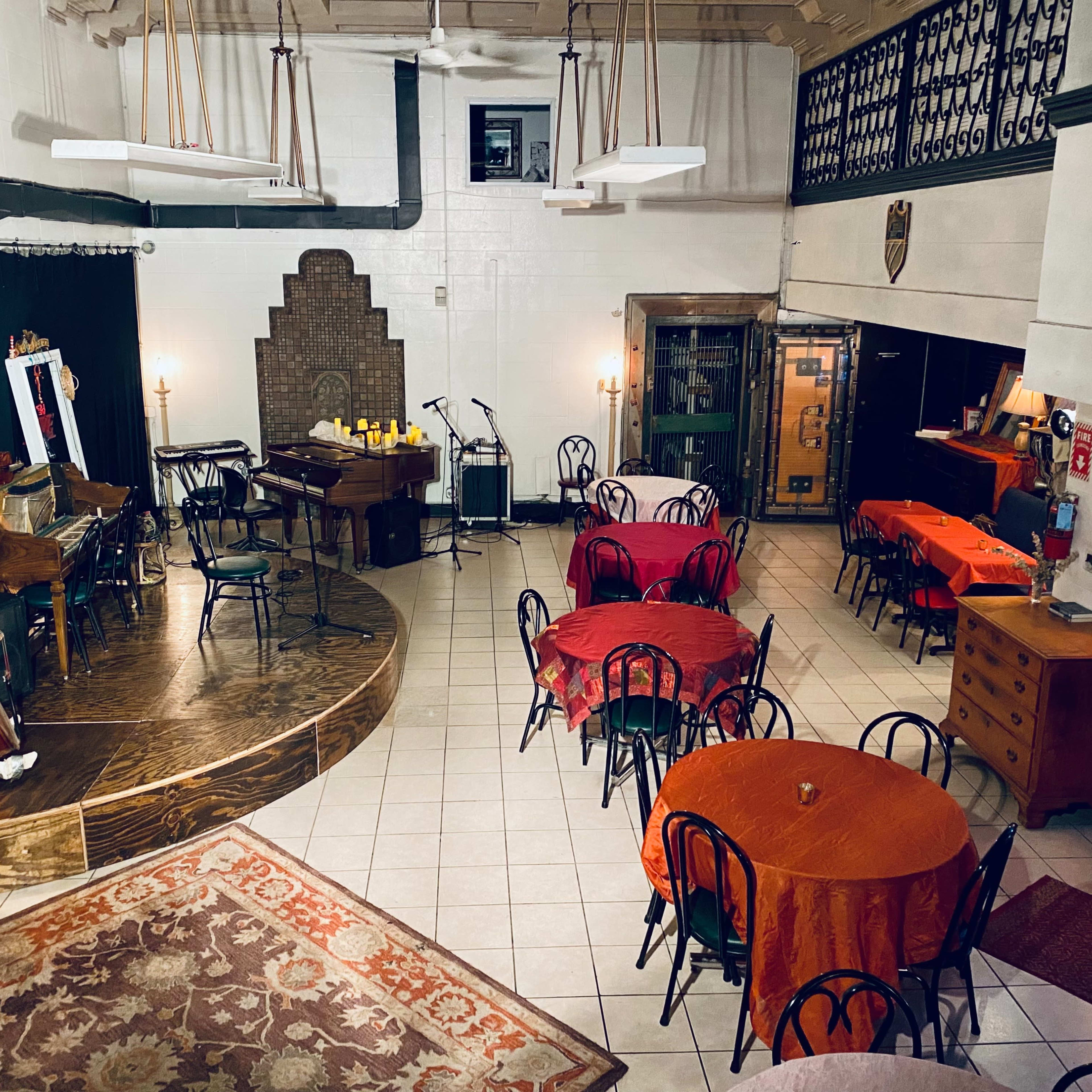 A spacious performance venue with a wooden stage, musical instruments, and several round tables set with orange tablecloths.
