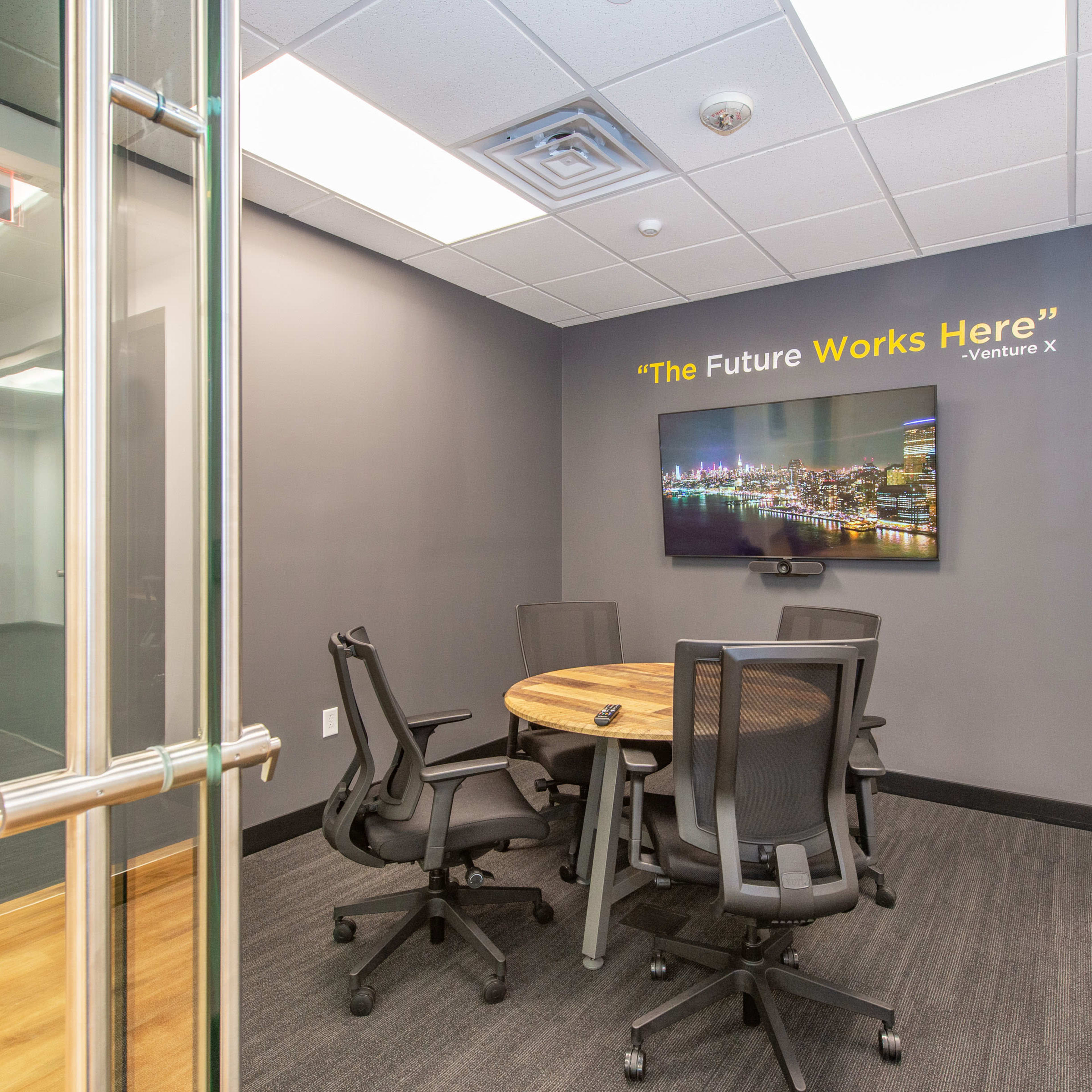 A modern conference room features a round wooden table surrounded by black chairs, with a television displaying the text "The Future Works Here" on the wall.