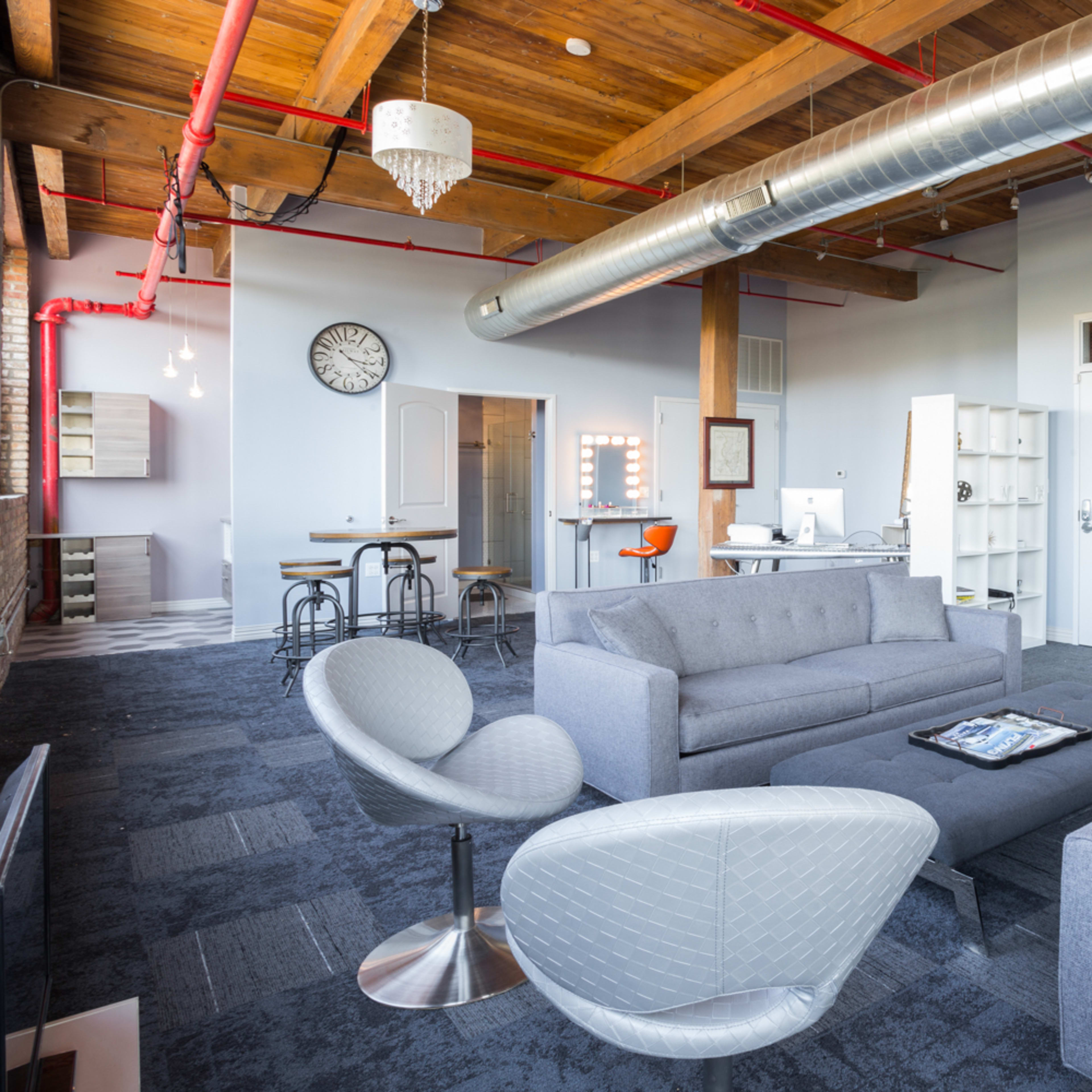 A modern office space featuring gray sofas, a low coffee table, and exposed wooden beams with industrial-style ventilation.