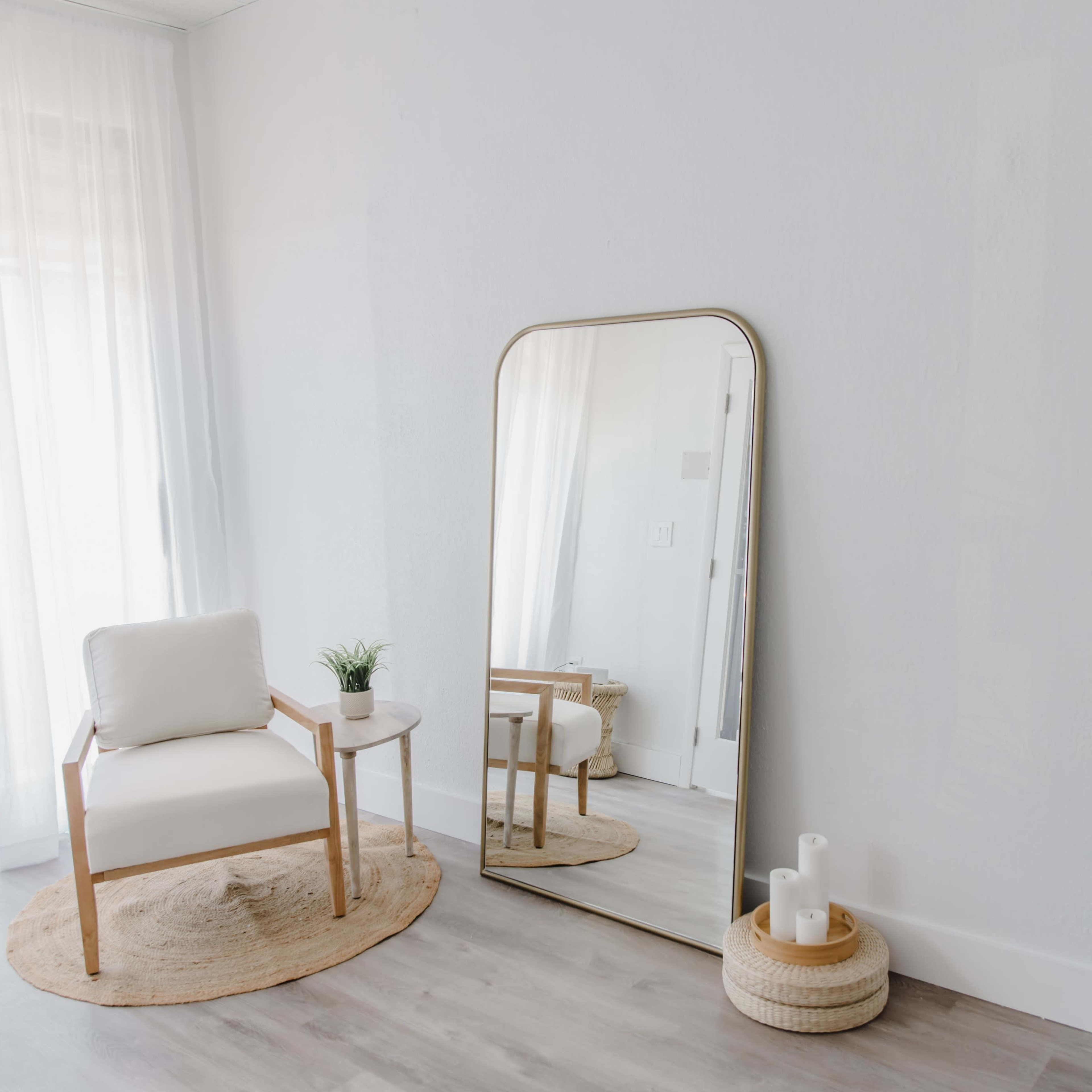 A minimalist interior features a white chair beside a round rug, a tall mirror reflecting light, and a small table with a potted plant.