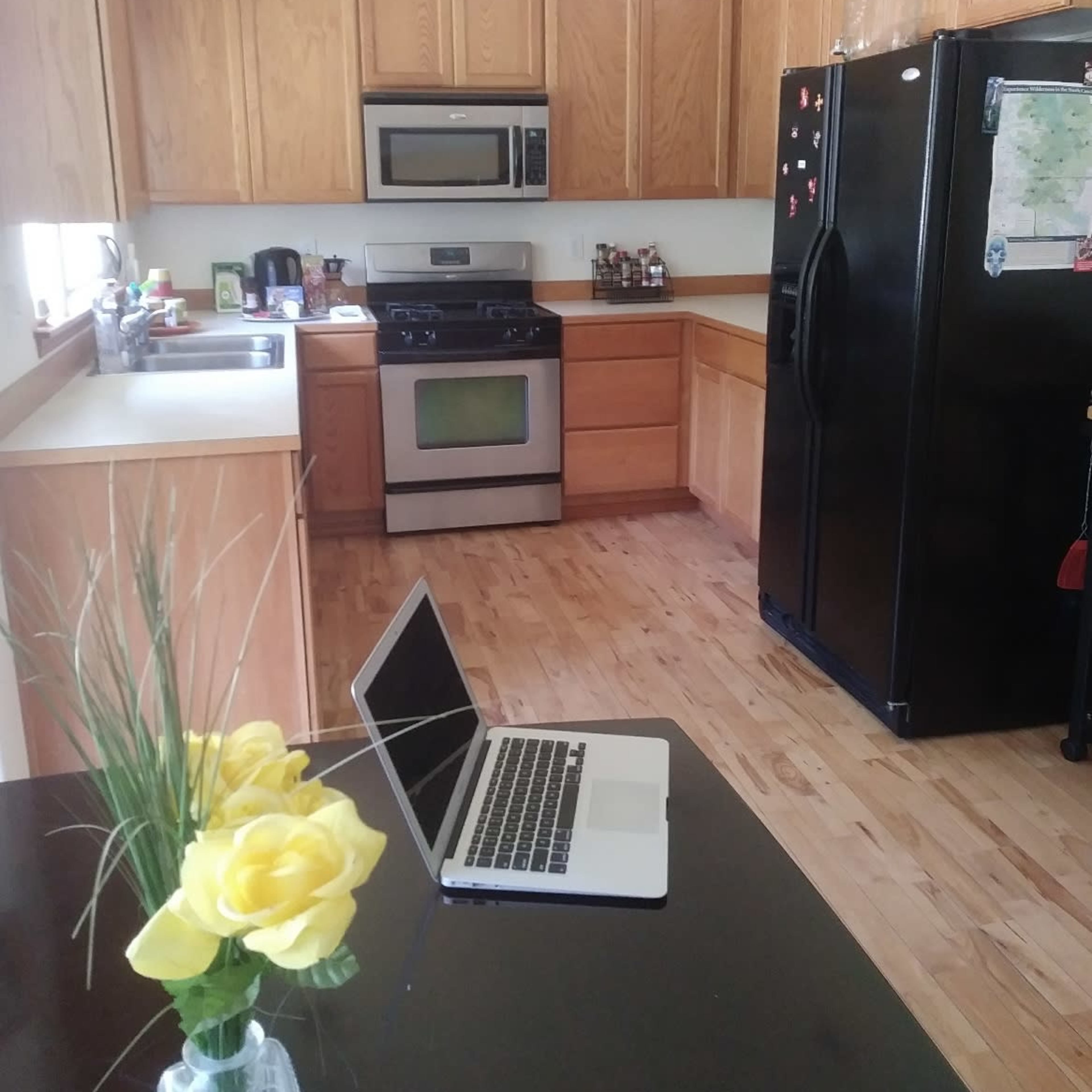 A kitchen with wooden cabinetry, a stainless steel microwave and oven, a black refrigerator, and a table with a laptop and a vase of yellow roses.