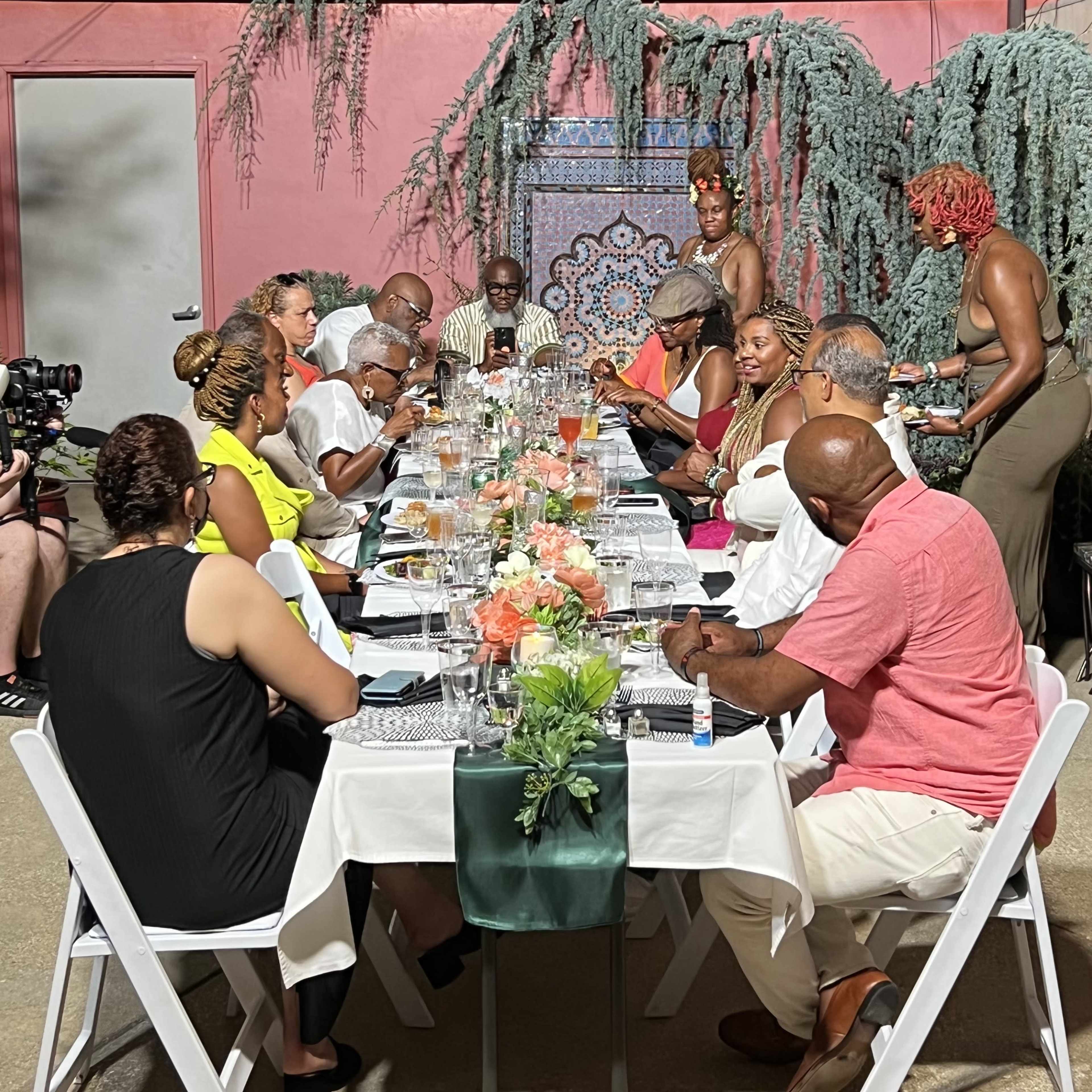 A group of people gathers around a long table set for a meal, adorned with glasses and floral centerpieces, while others take photos and engage in conversation.