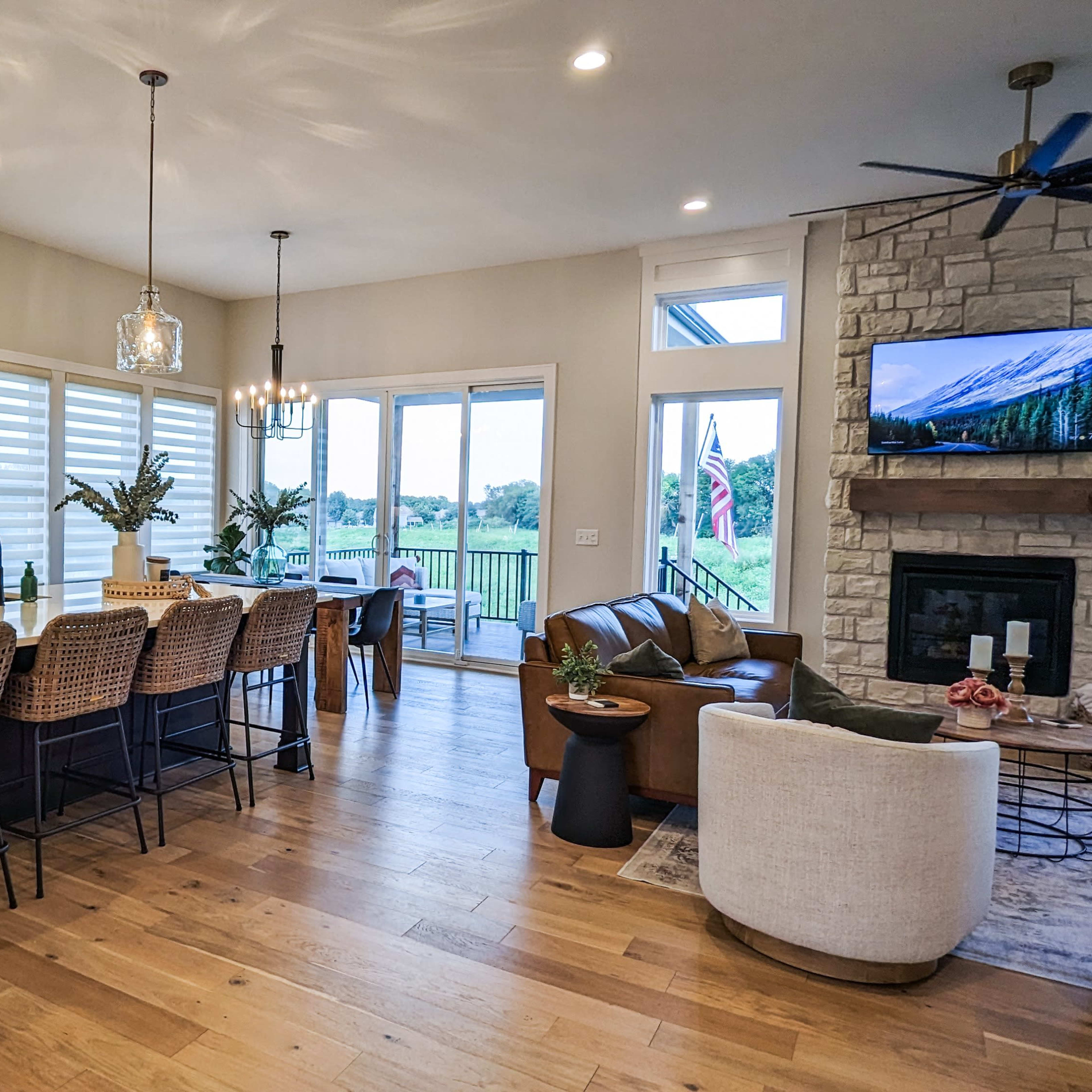 A modern open-concept kitchen and living area features a large island with bar stools, stone accents, and expansive windows overlooking a scenic view.