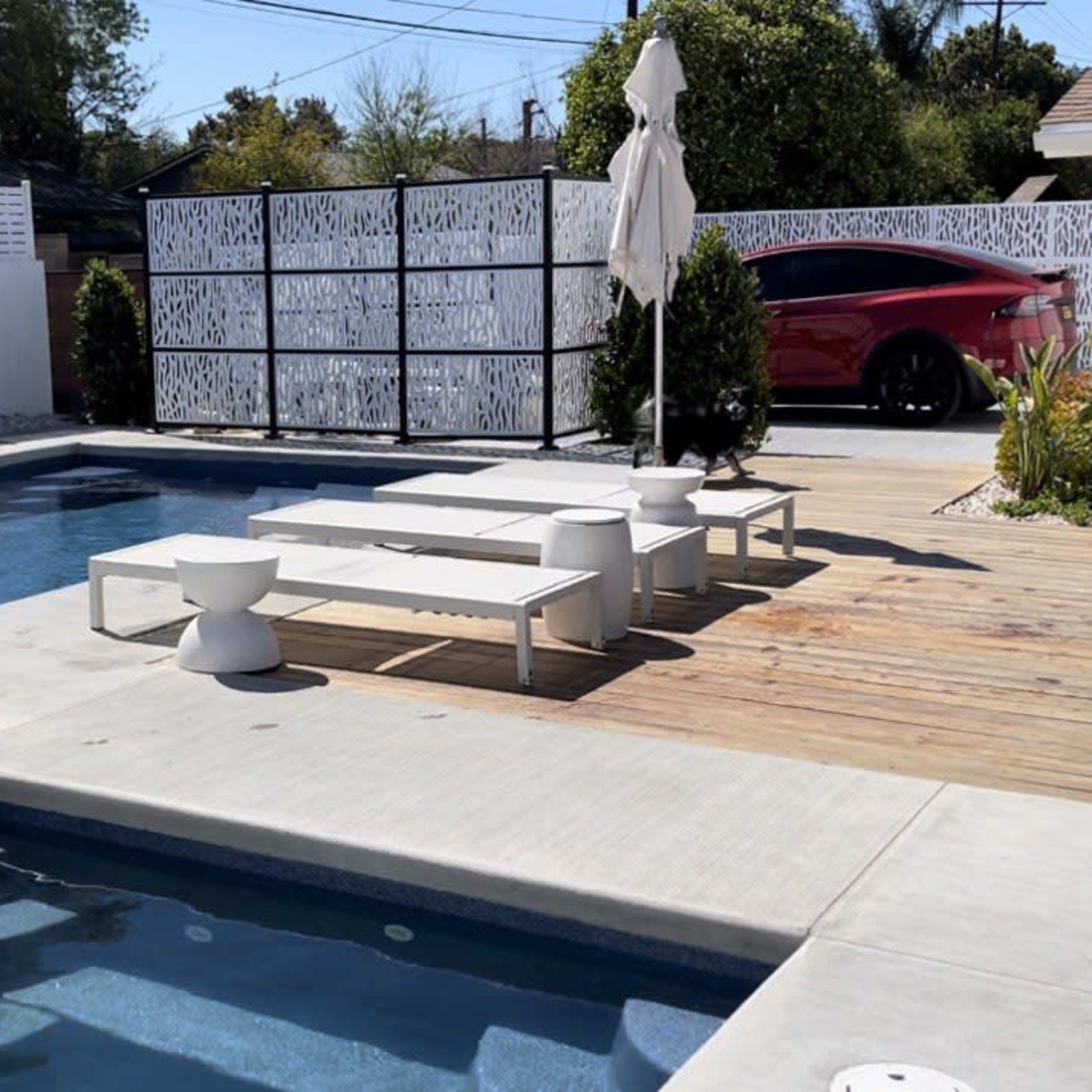 A modern backyard pool area featuring a large swimming pool, a wooden deck with white outdoor furniture, and a sleek car parked in the background.