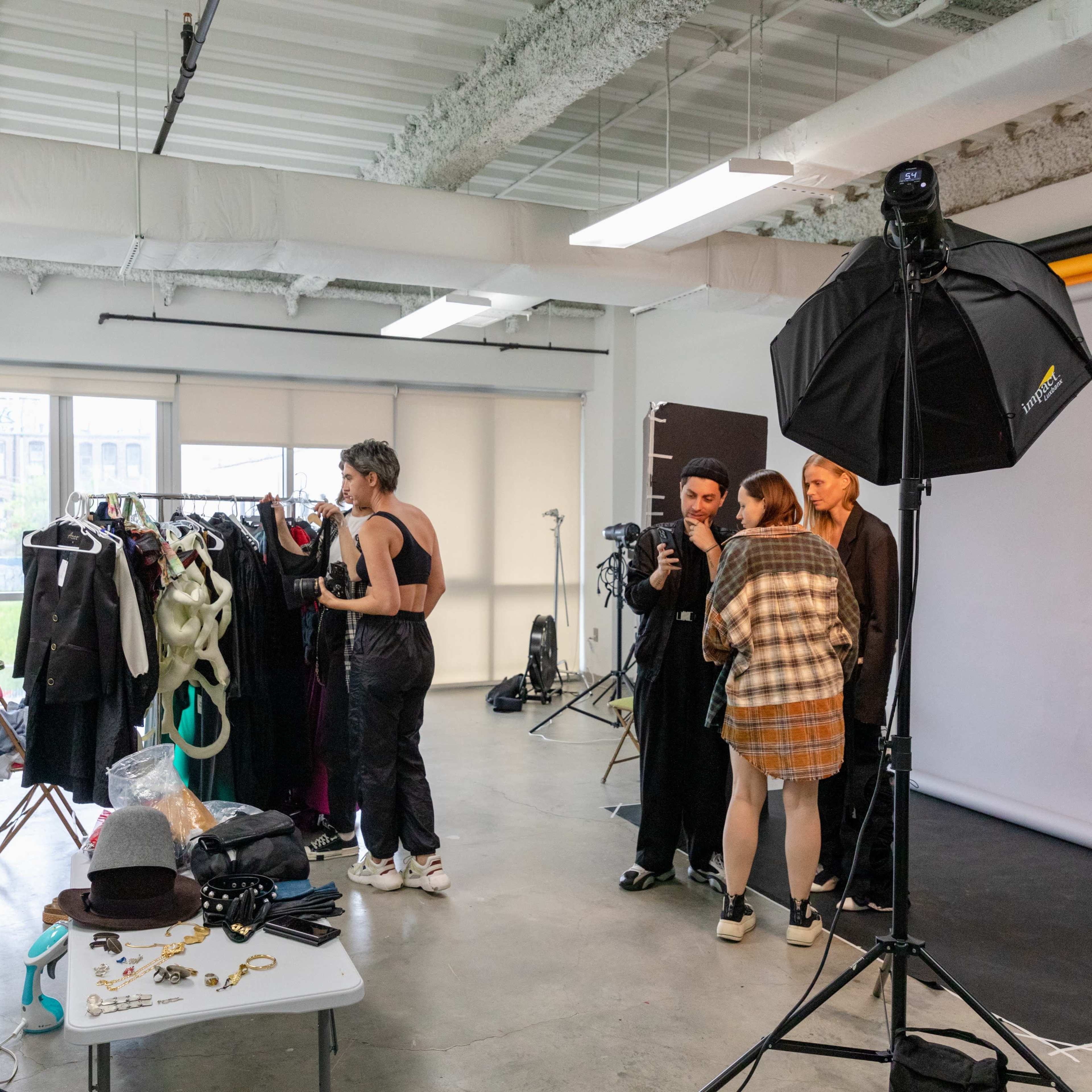 A group of people is gathered in a photography studio with clothing racks, equipment, and a backdrop.