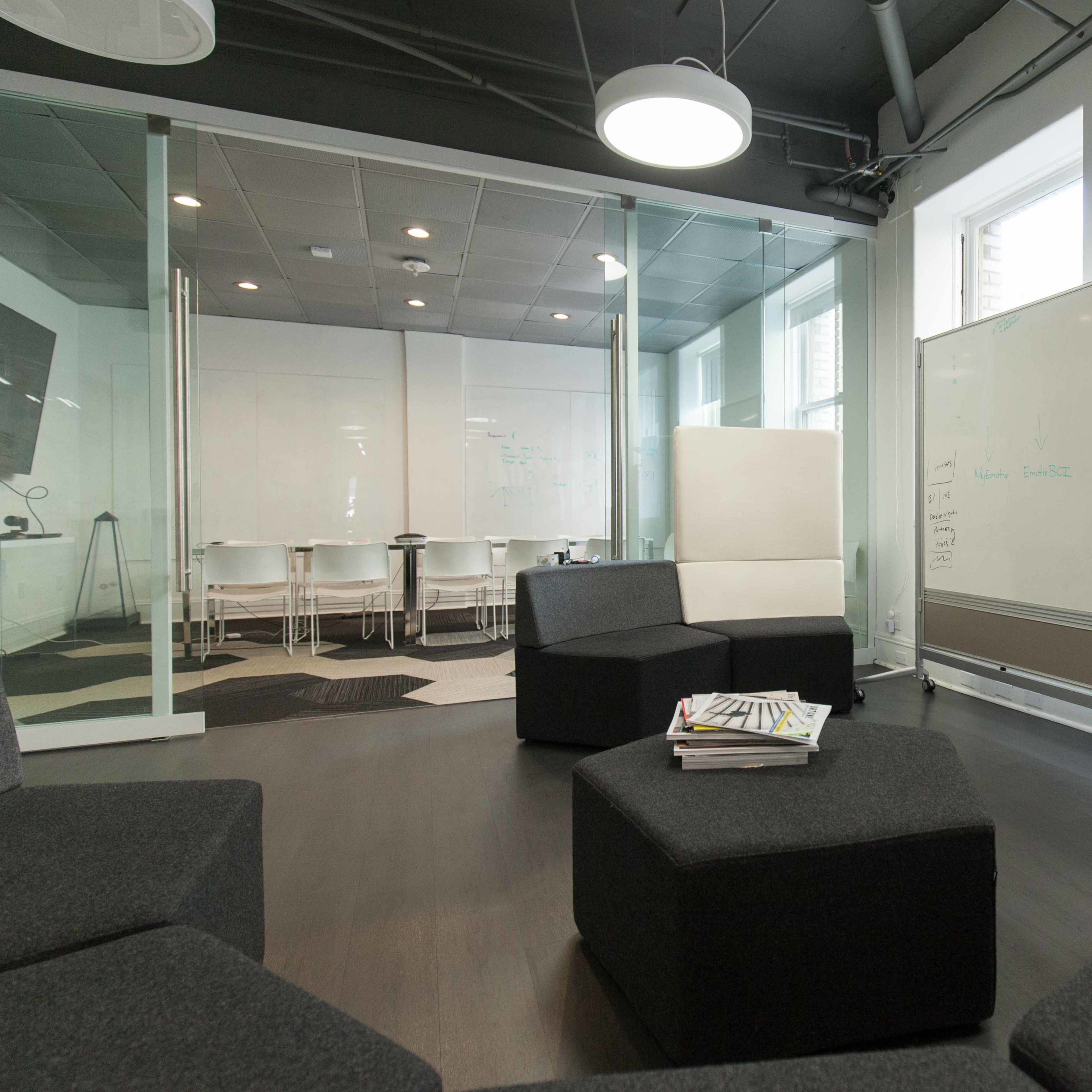 A modern office space featuring a seating area with modular black and gray furniture and a glass-walled meeting room in the background.