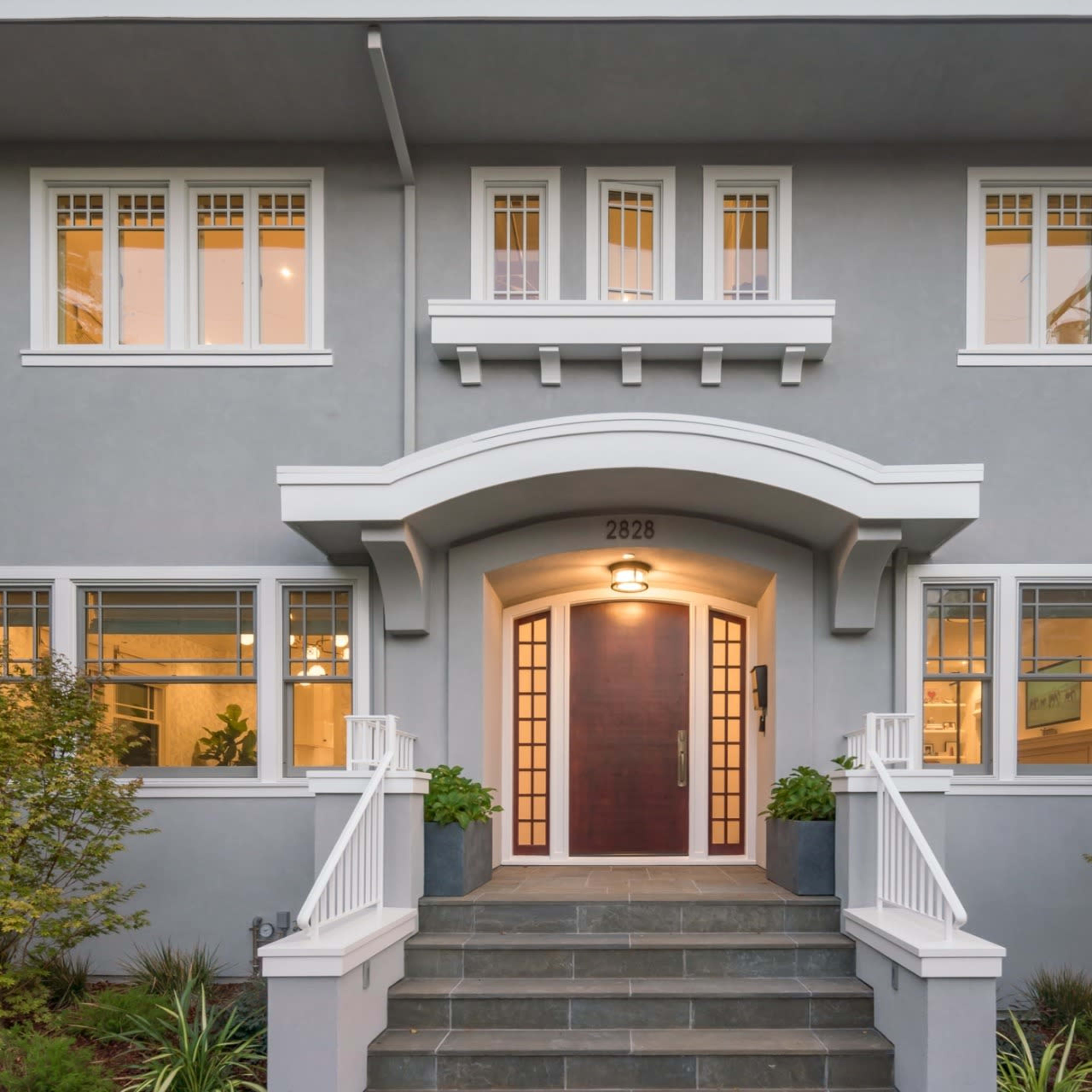 A modern two-story house with a gray exterior, large front windows, and a central wooden door framed by a white archway and stairway.
