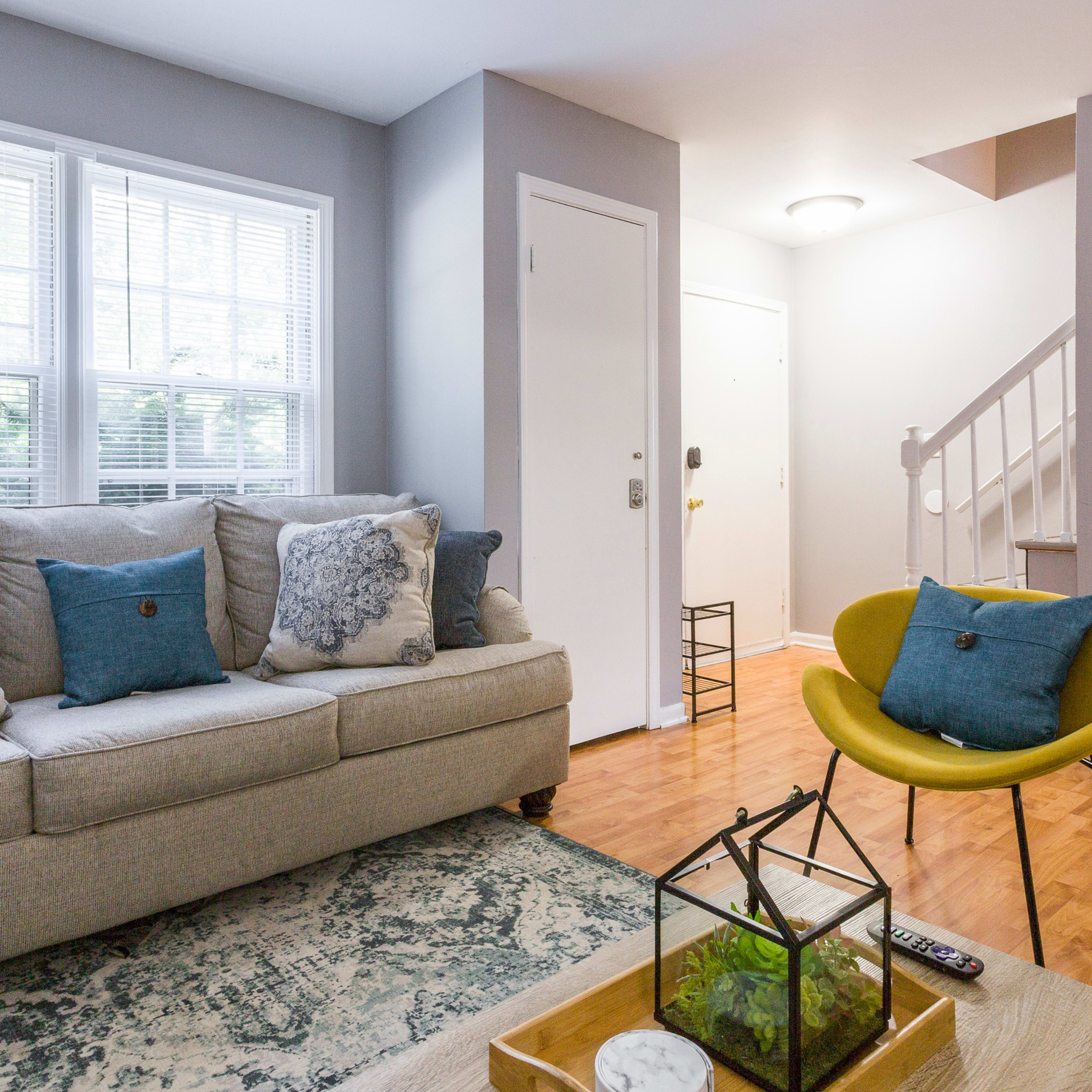 A gray sofa with decorative pillows sits in a living room beside a glass coffee table, facing a staircase and an entrance door.