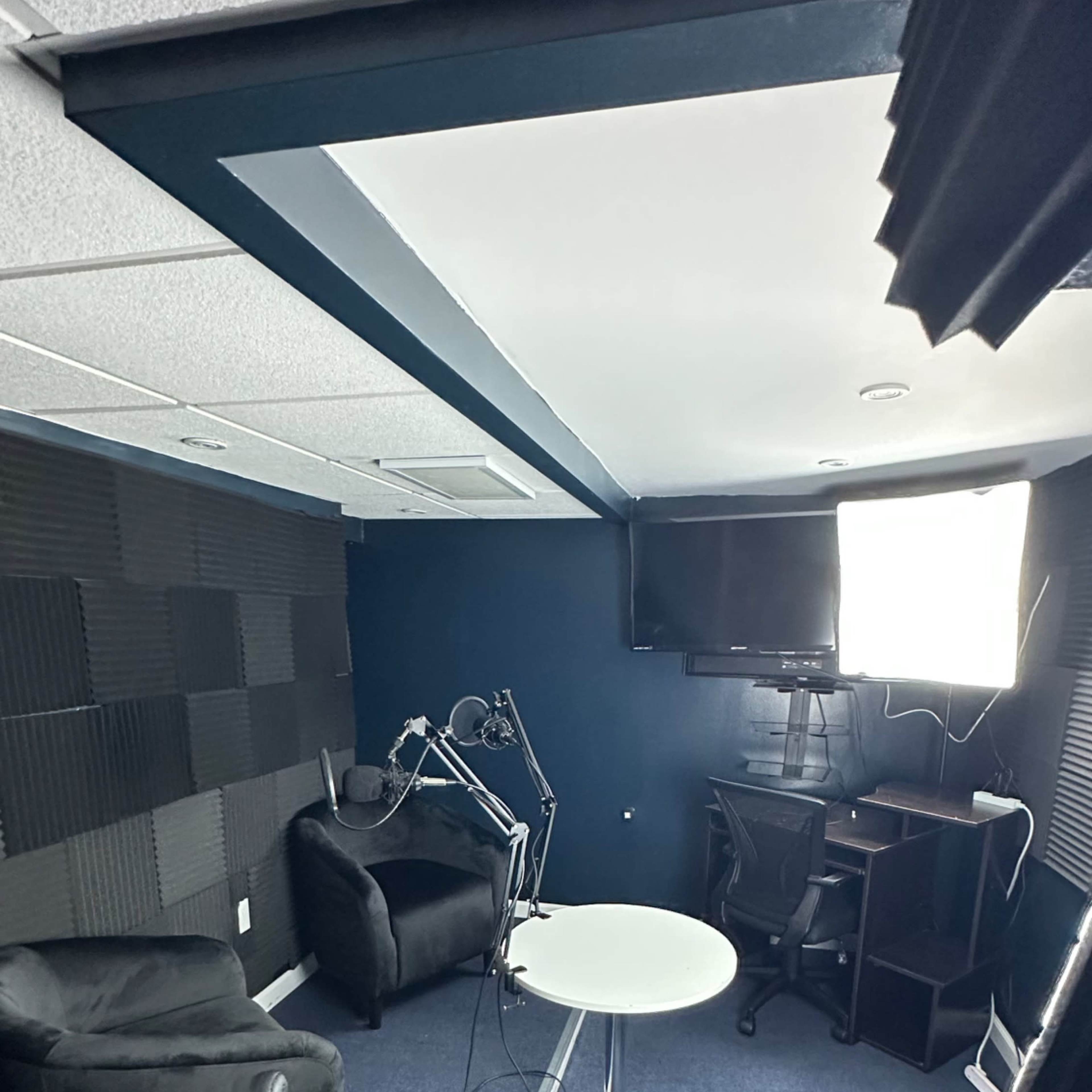 A small, dimly lit recording studio with soundproof walls, a table in the center, and black furniture arranged around it.