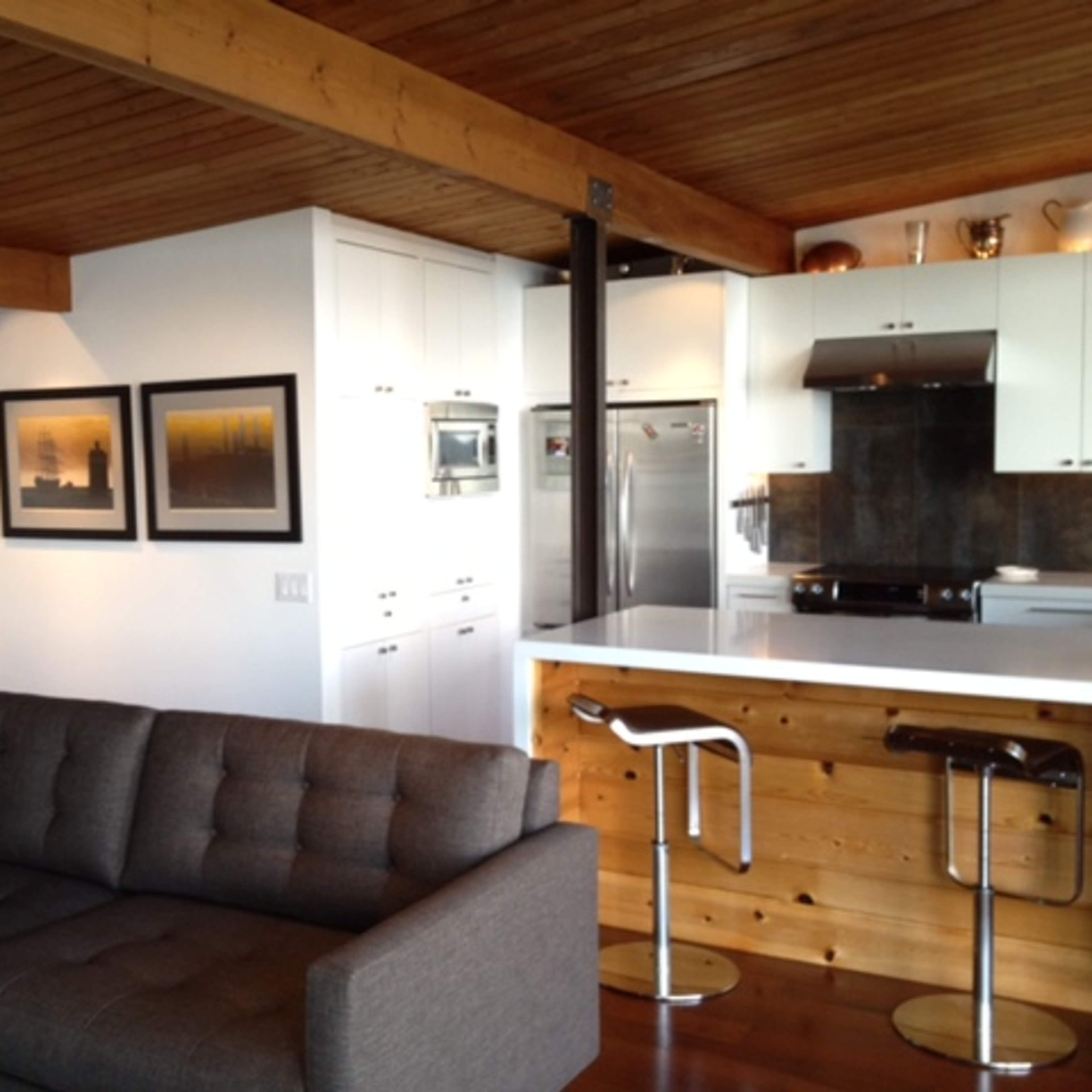 A modern open-plan living area with a gray sofa, wooden ceiling beams, and a kitchen featuring white cabinetry and stainless steel appliances.