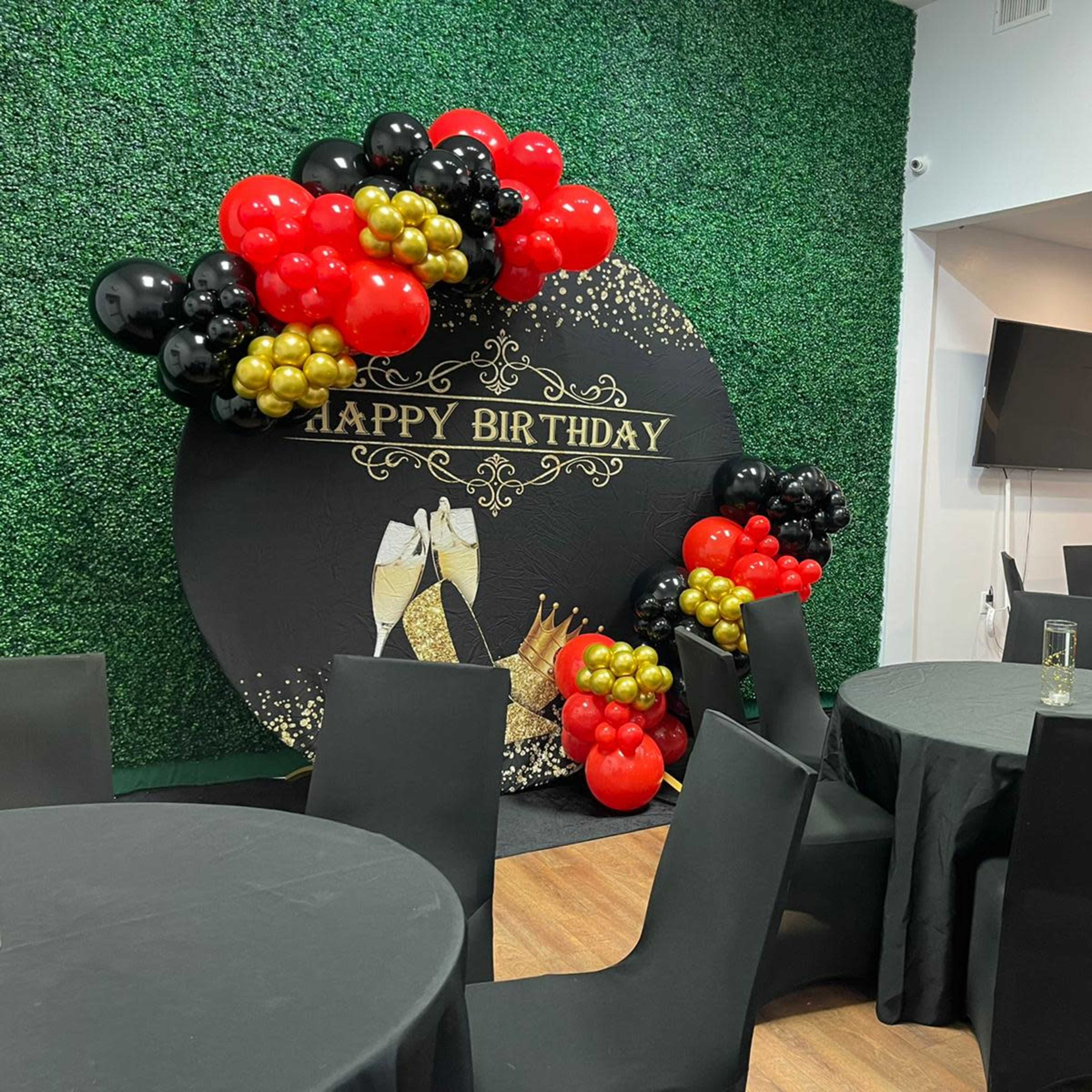 A birthday party decoration featuring a green wall adorned with a black and gold "Happy Birthday" sign, surrounded by clustered balloons in red, black, and gold, with several tables set up in front.
