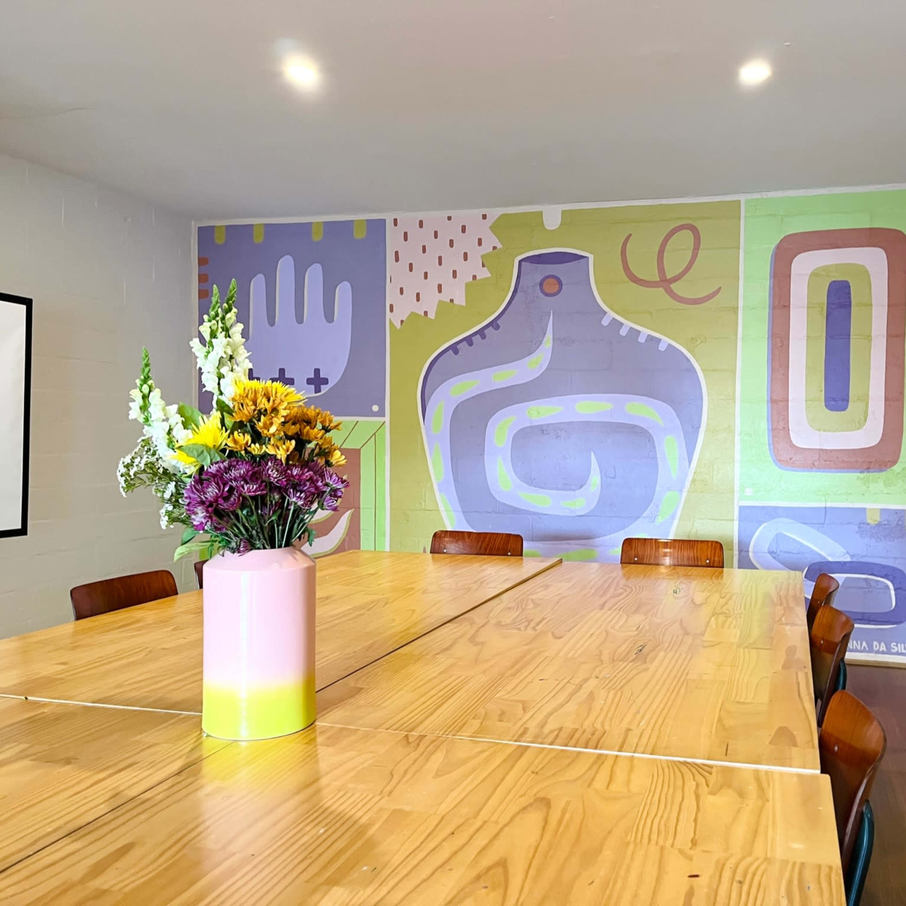 A wooden table surrounded by chairs sits in a room with a colorful mural on the wall and a flower arrangement in the center.