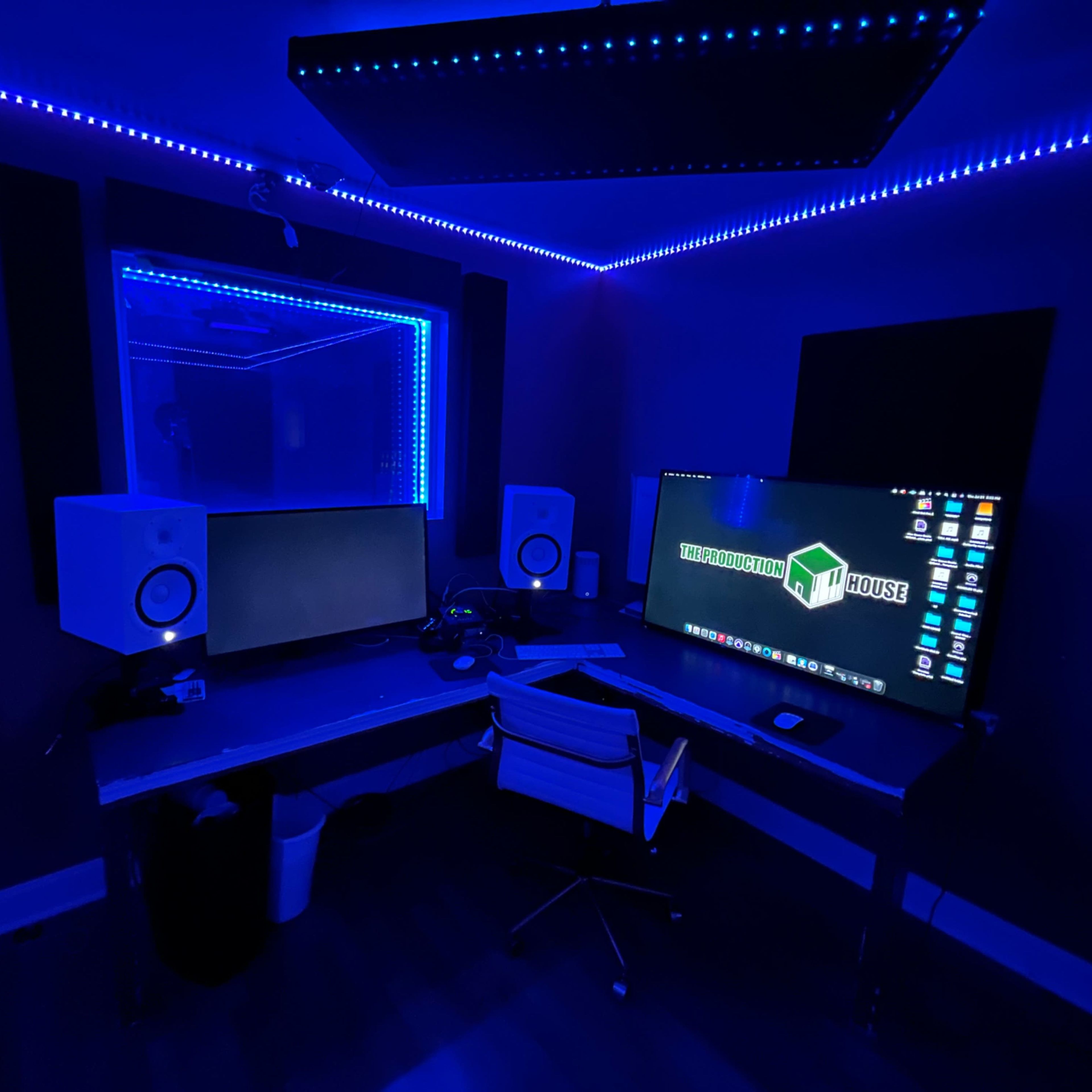 A dimly lit recording studio features blue LED strip lighting, two monitors, a pair of speakers, and a sleek desk with a rolling chair.