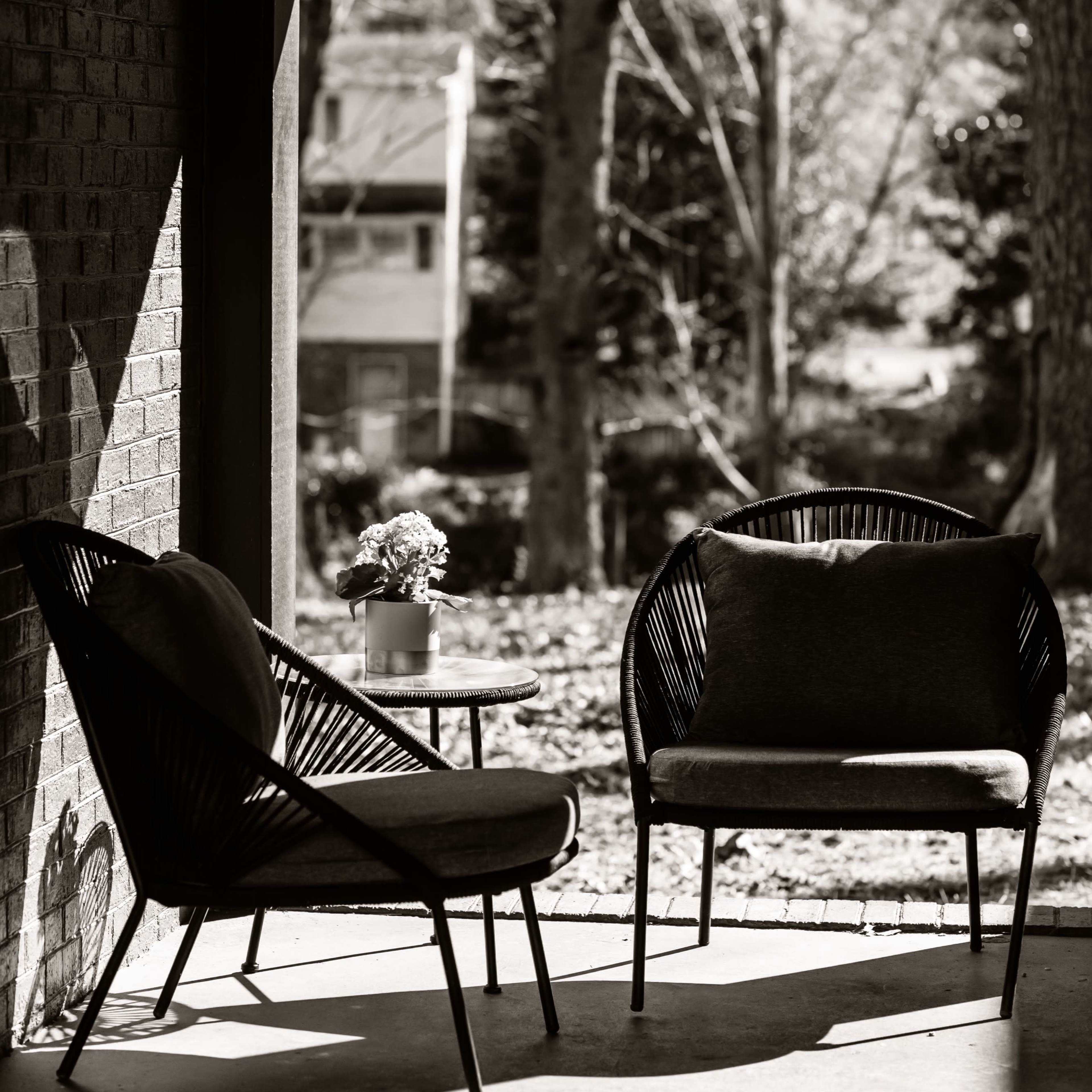 Two black rattan chairs and a small round table with a potted plant, framed by a shaded porch overlooking a wooded area.