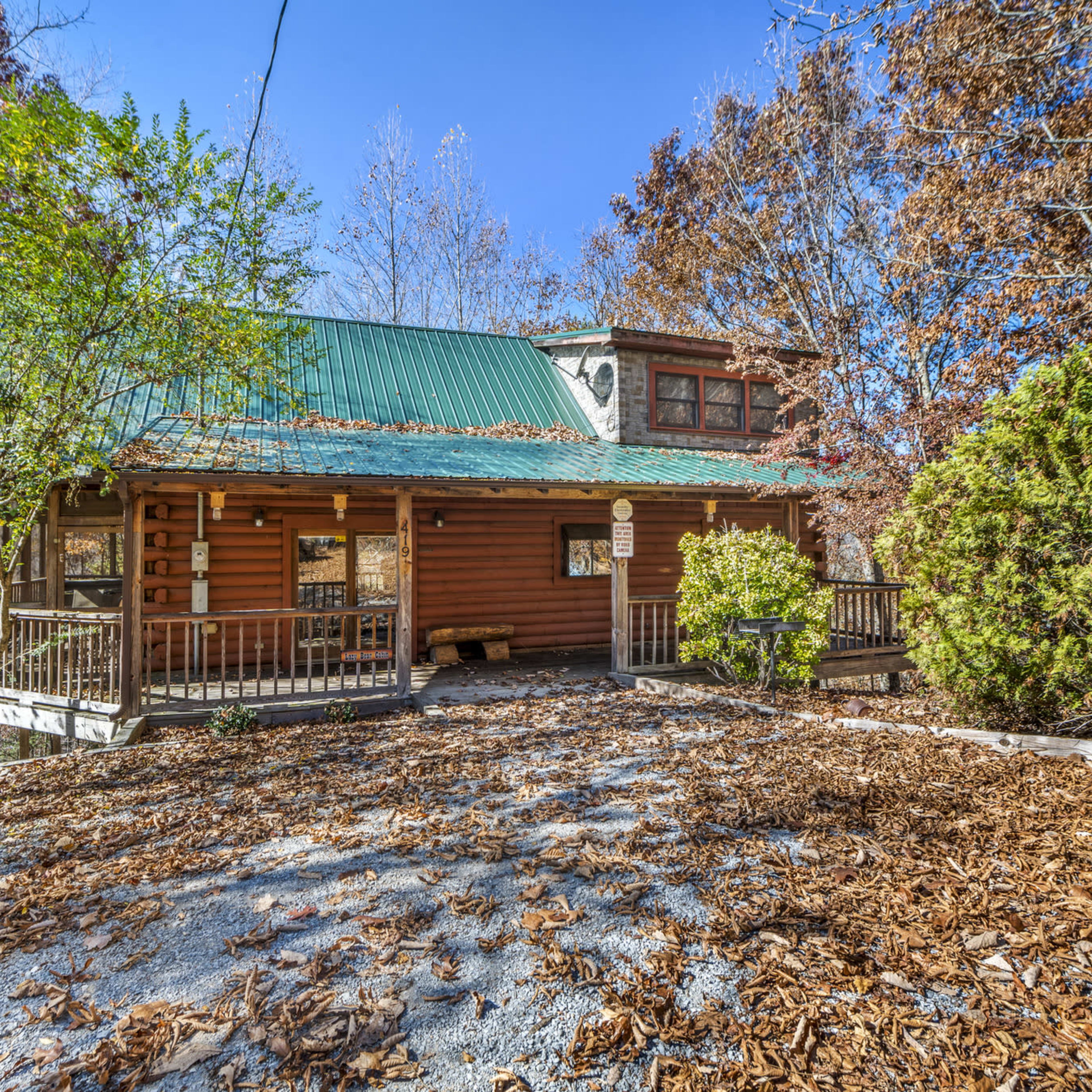 A wooden cabin with a green metal roof is surrounded by trees and a layer of dry leaves on the ground.