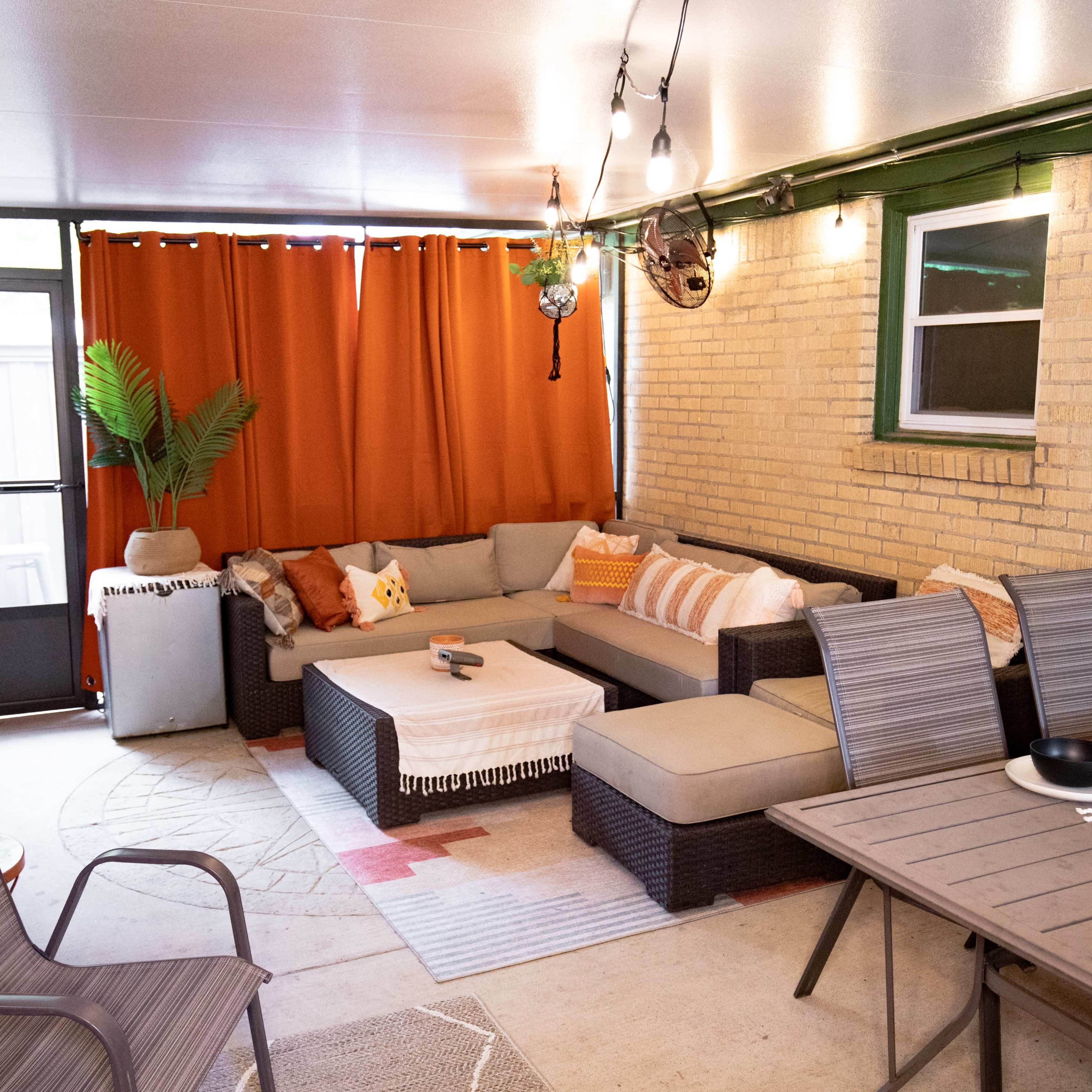 A cozy, covered patio area featuring a sectional sofa, dining table, and decorative plants, with orange curtains and string lights accentuating the space.