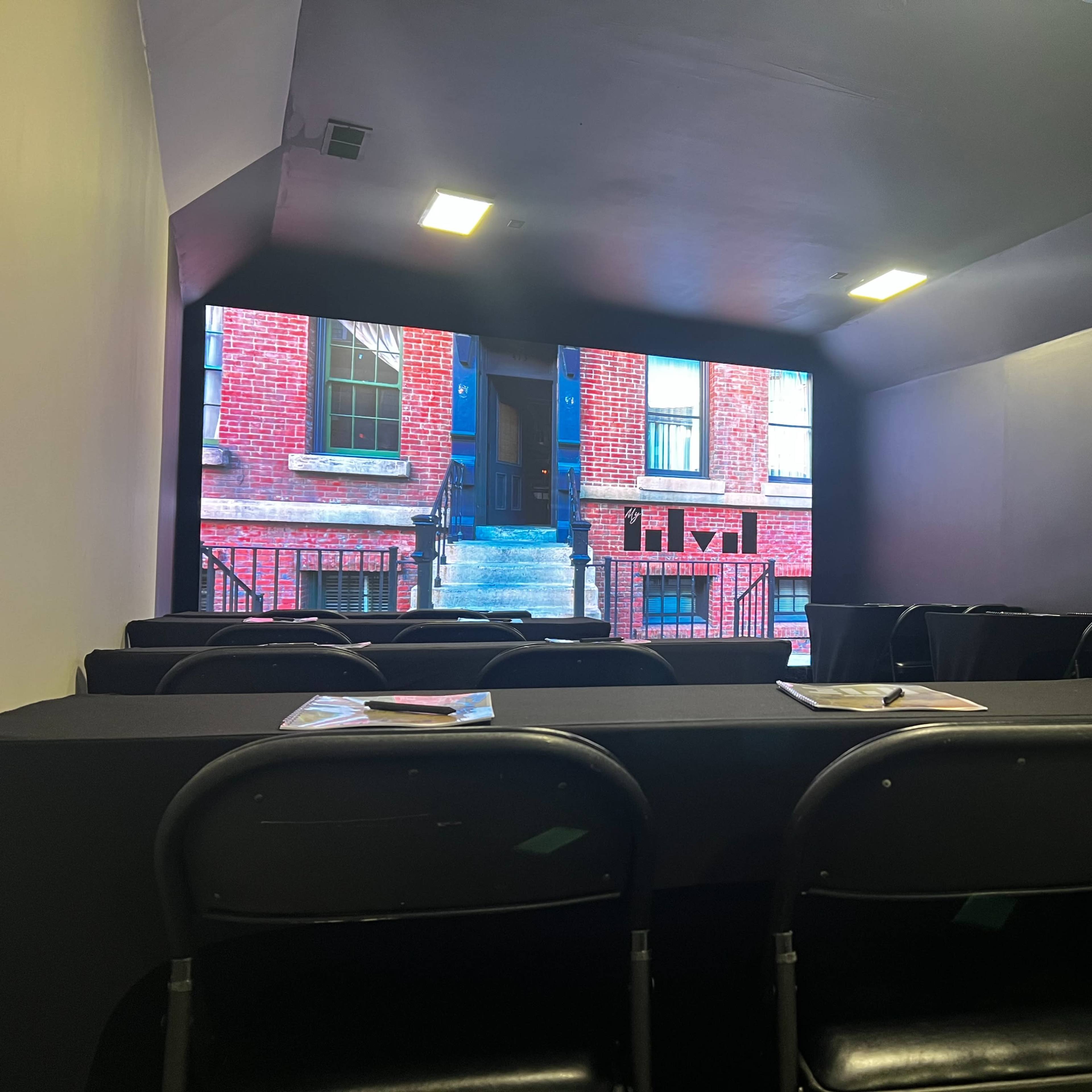 A small presentation room featuring a large screen displaying a brick building, with several black folding chairs arranged in front of a table.