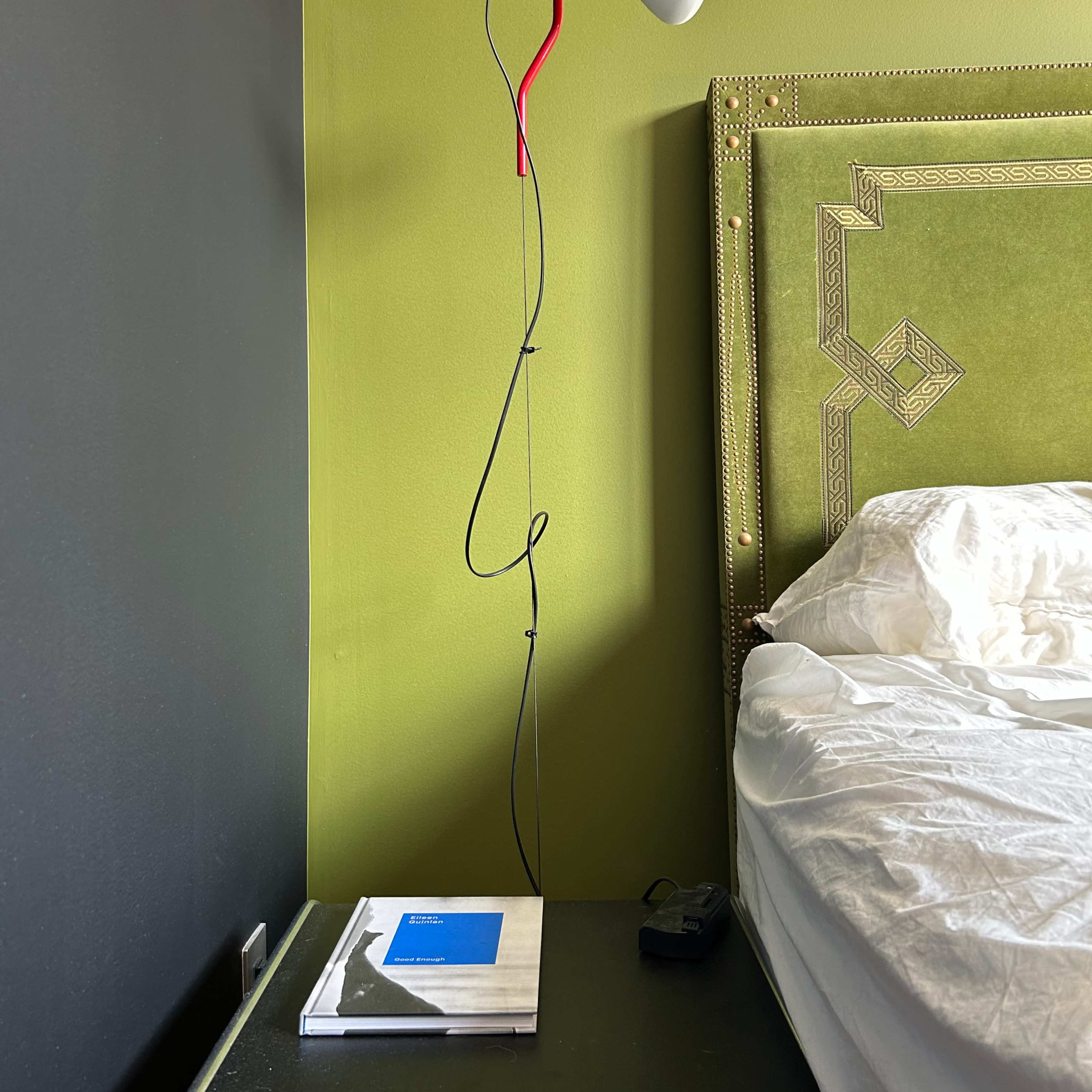 A black bedside table with a stack of books sits next to a bed with rumpled white bedding, while a lamp with a white shade and red cord stands against a green wall.