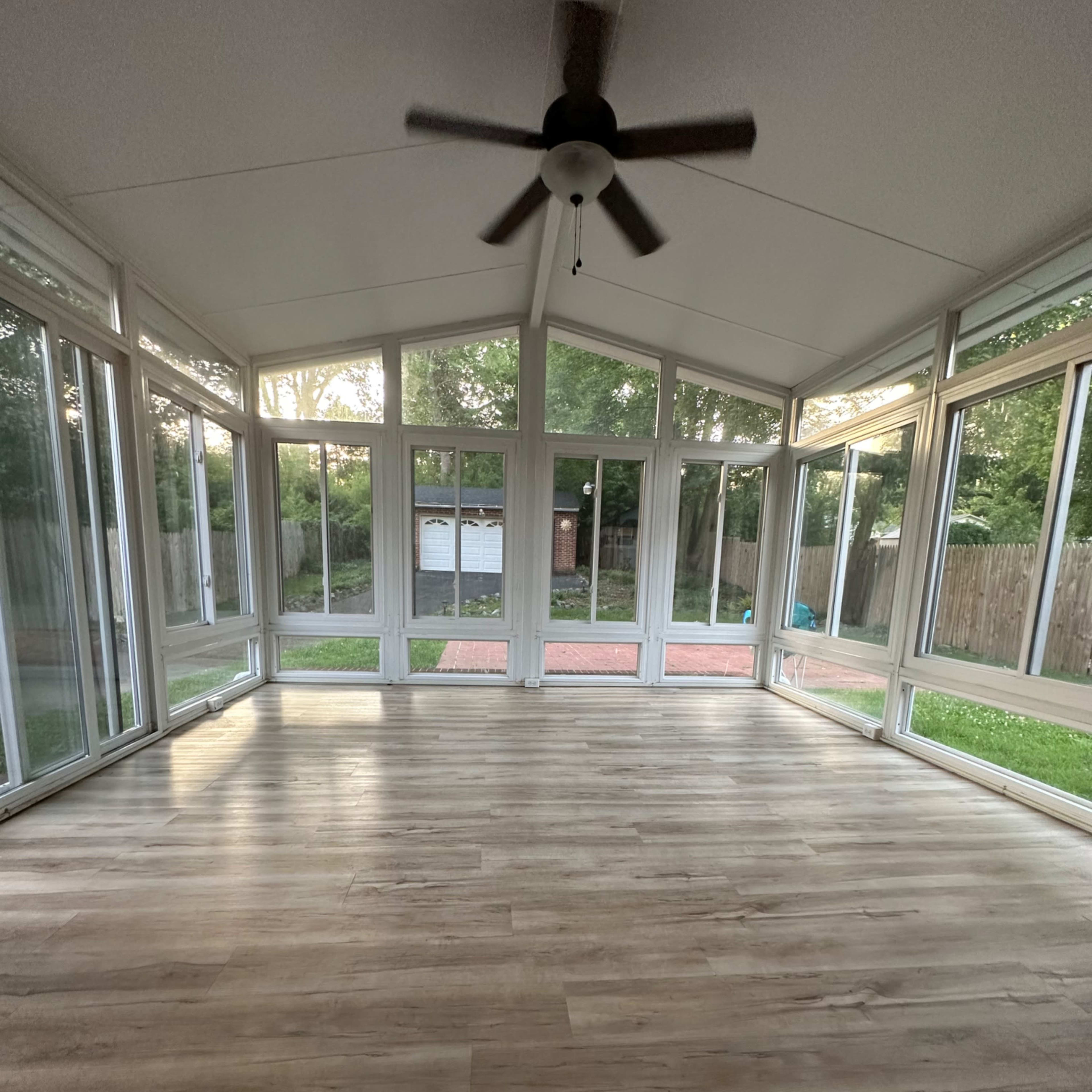 A spacious sunroom with large windows, a ceiling fan, and laminate flooring, overlooking a grassy area and a partially visible outdoor space.