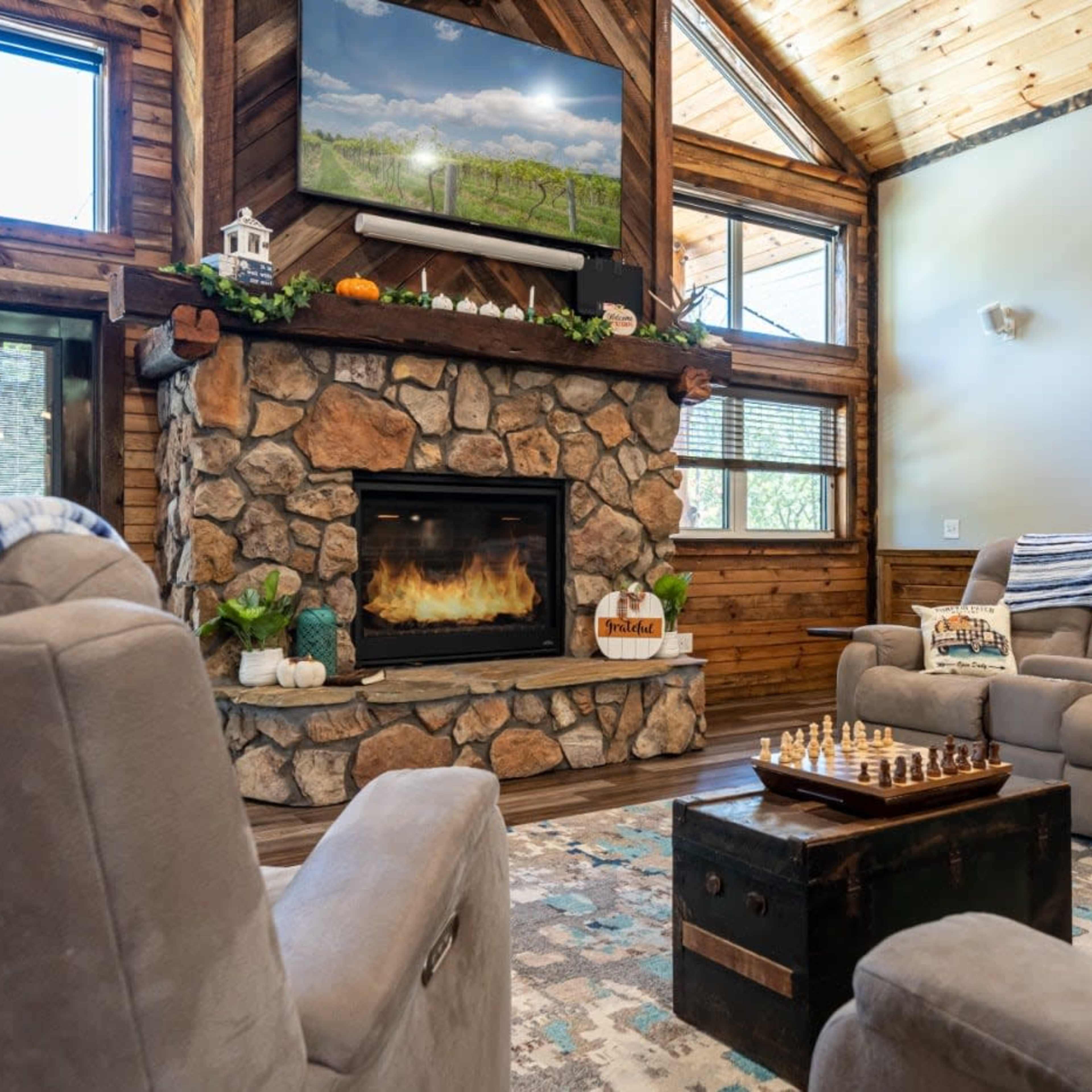 A cozy living room featuring a stone fireplace, a mounted deer head, and a chess set on a small table, with wooden walls and a television mounted above the fireplace.