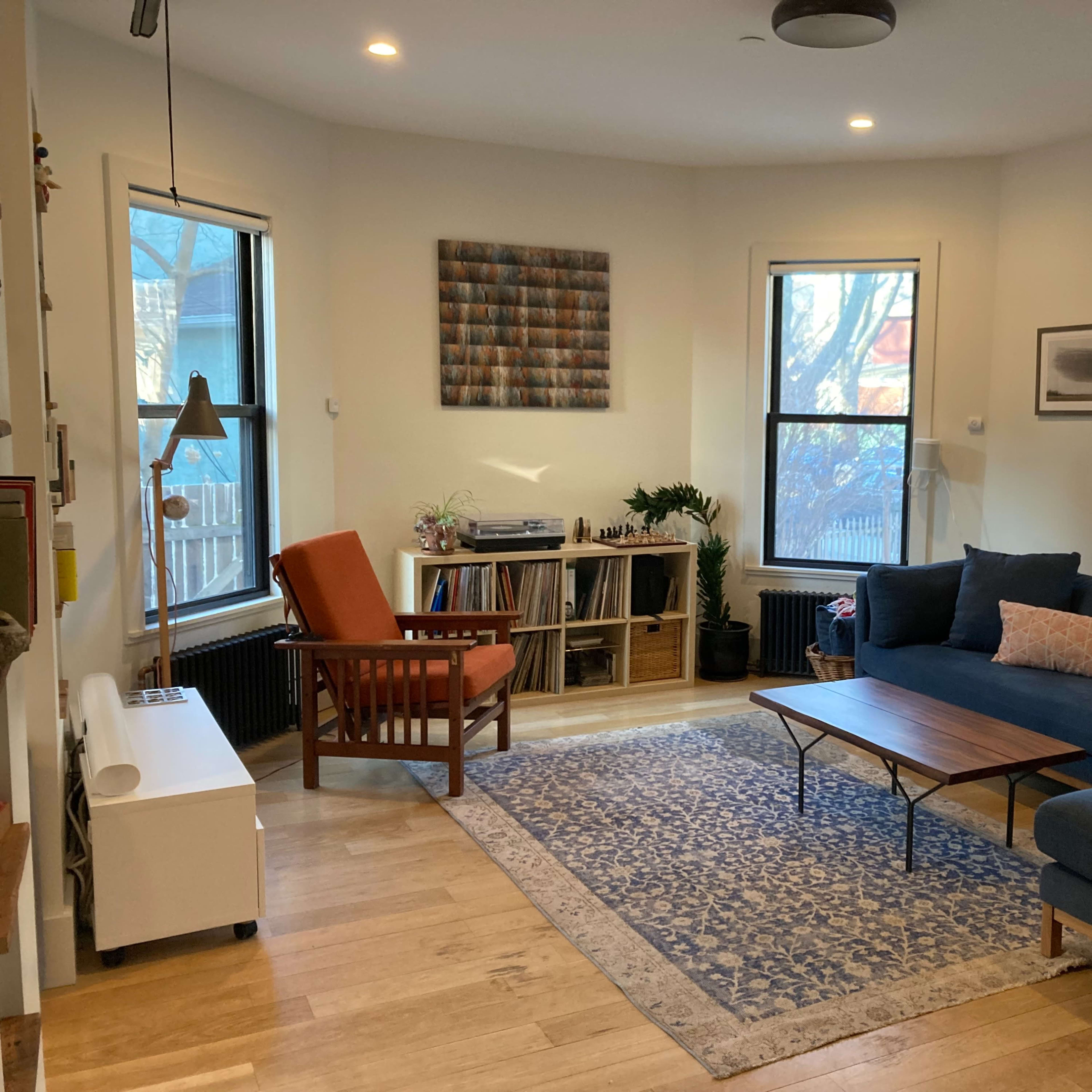 A cozy living room with a blue sofa, a wooden coffee table, a record player, and large windows letting in natural light.