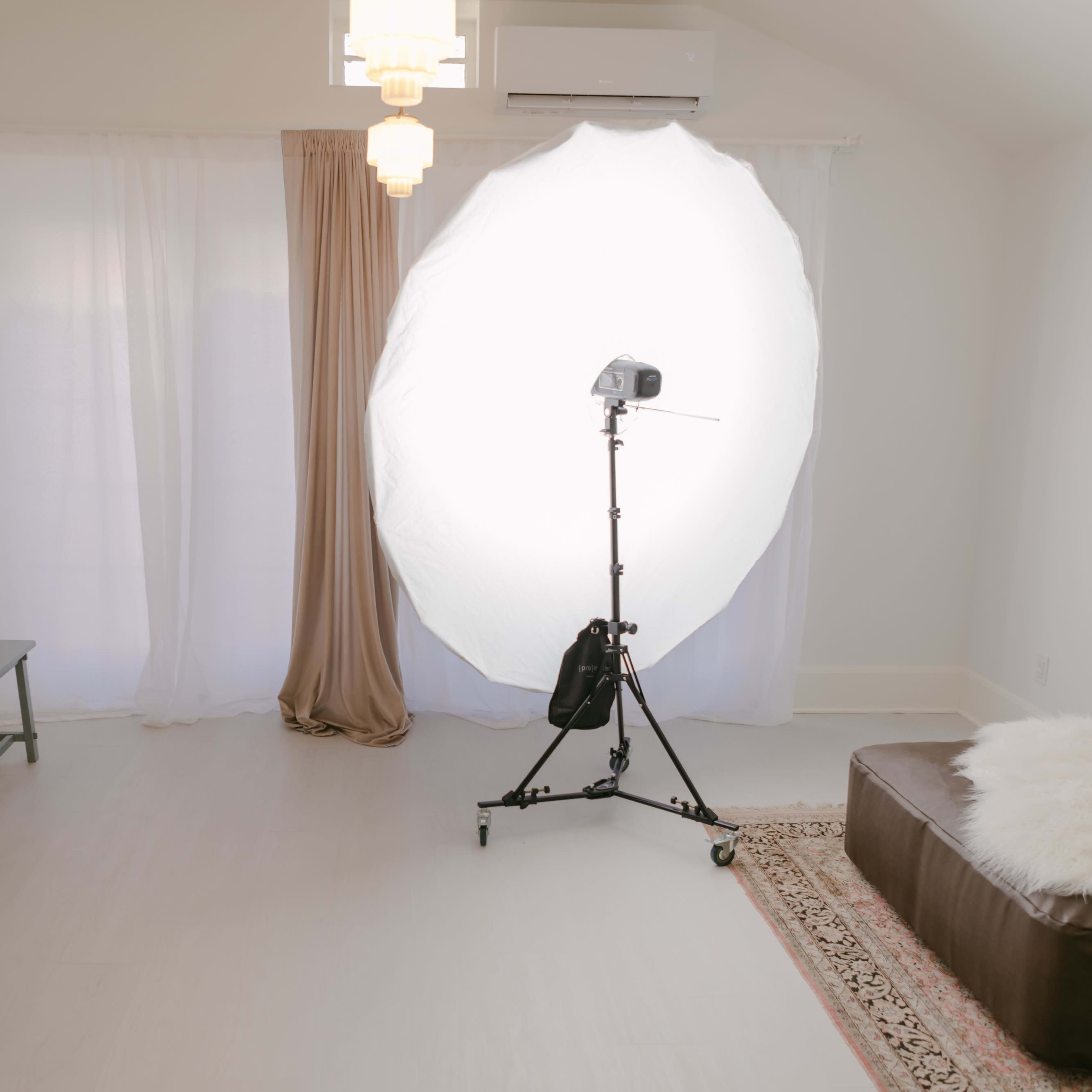 A bright, minimalistic interior space featuring a softbox lighting setup, a cream-colored sofa, a green table, and a brown leather ottoman with a white fur throw.
