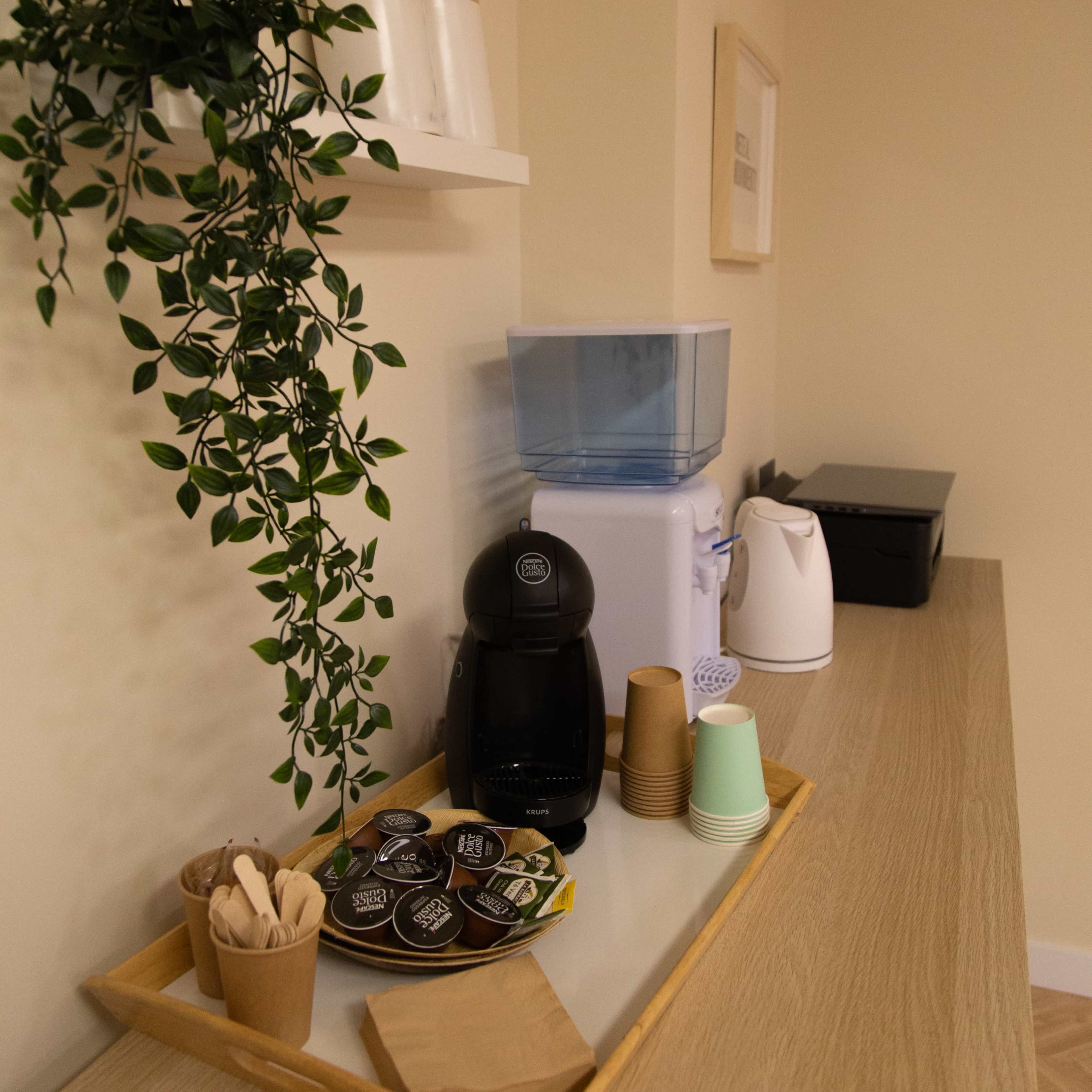 A countertop features a coffee maker, a water dispenser, a stack of paper cups, trays with snacks, and a small potted plant.