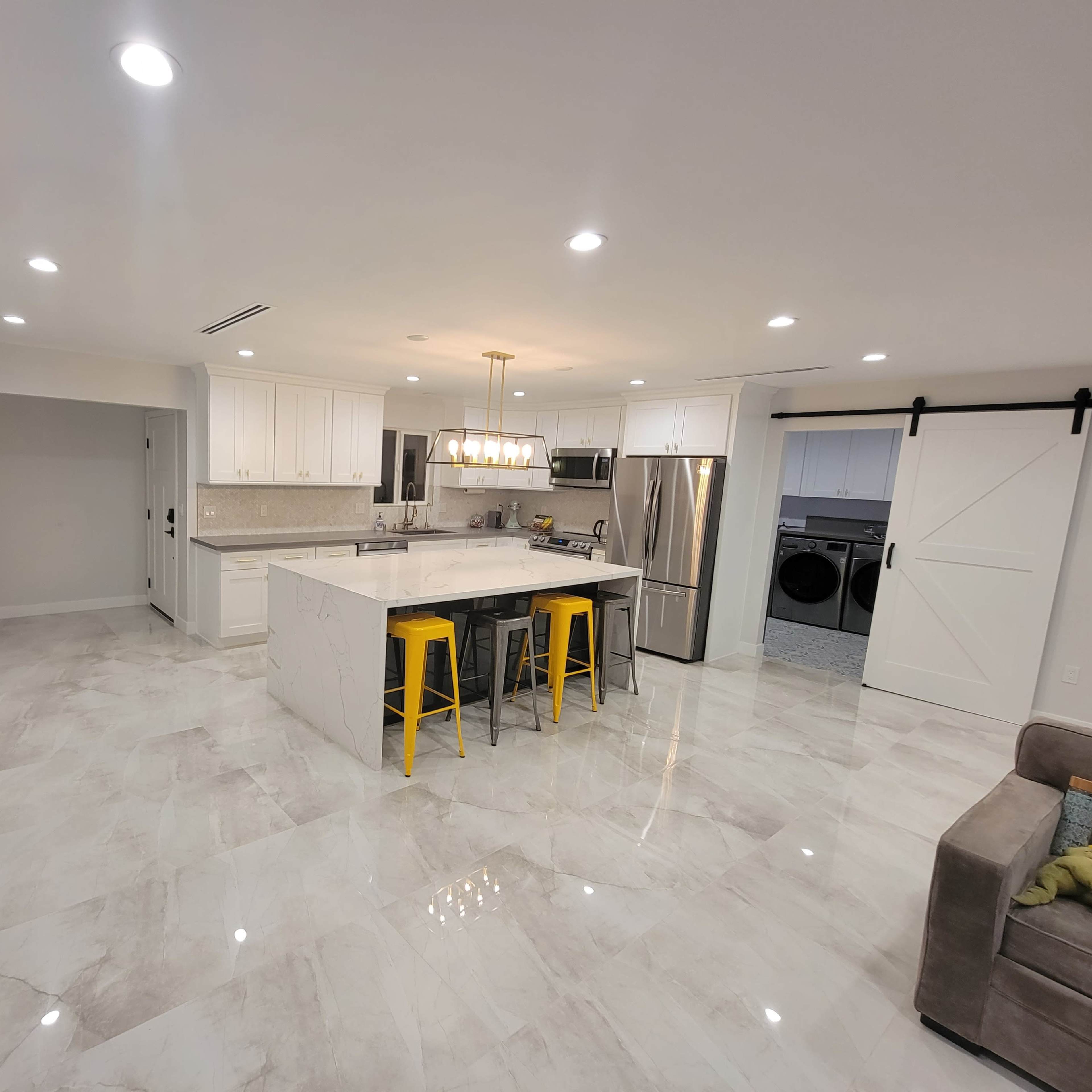 A modern kitchen features white cabinetry, a central island with yellow bar stools, stainless steel appliances, and polished marble flooring.
