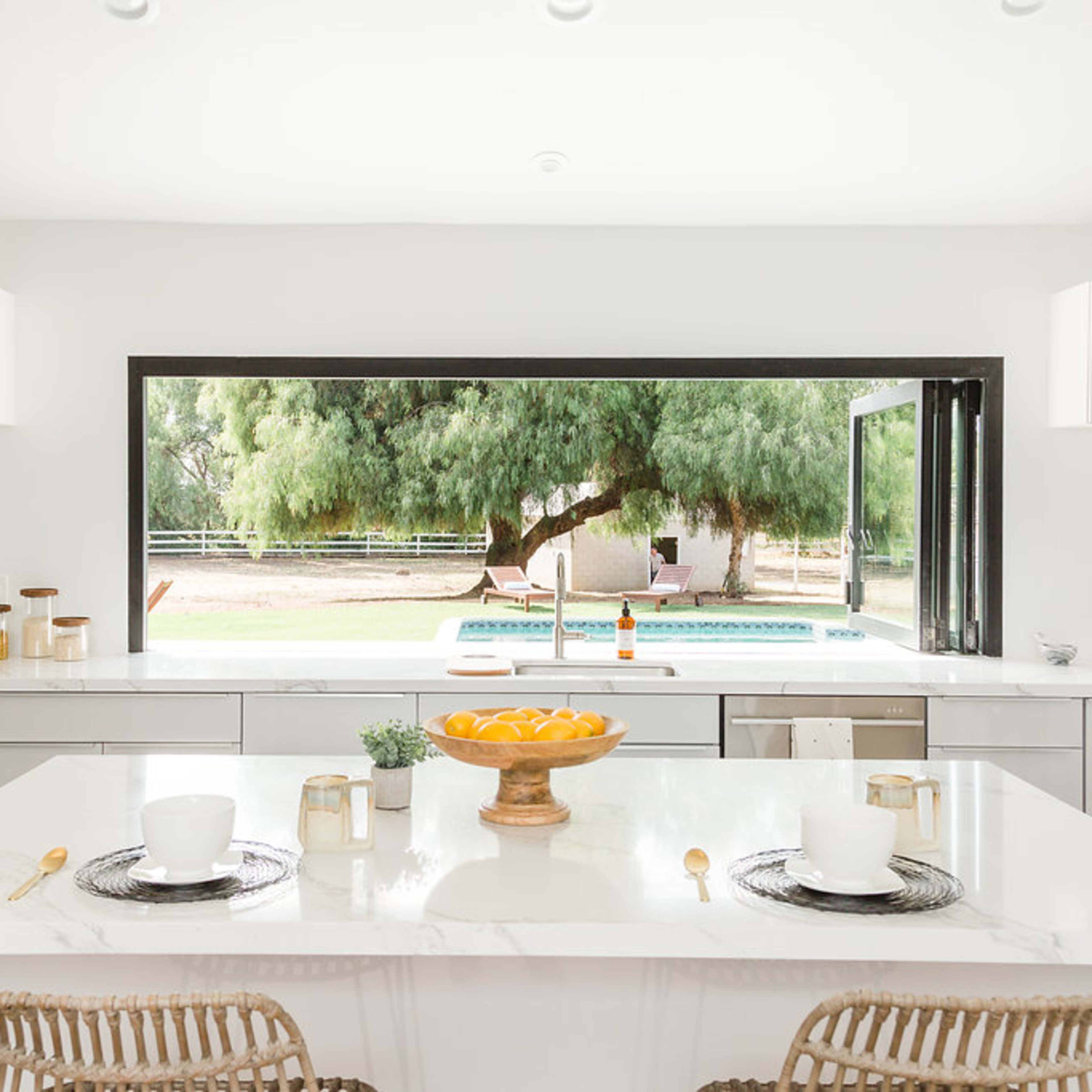 A modern kitchen features a large island with a bowl of fruit, overlooking a patio and lush greenery through a wide window.