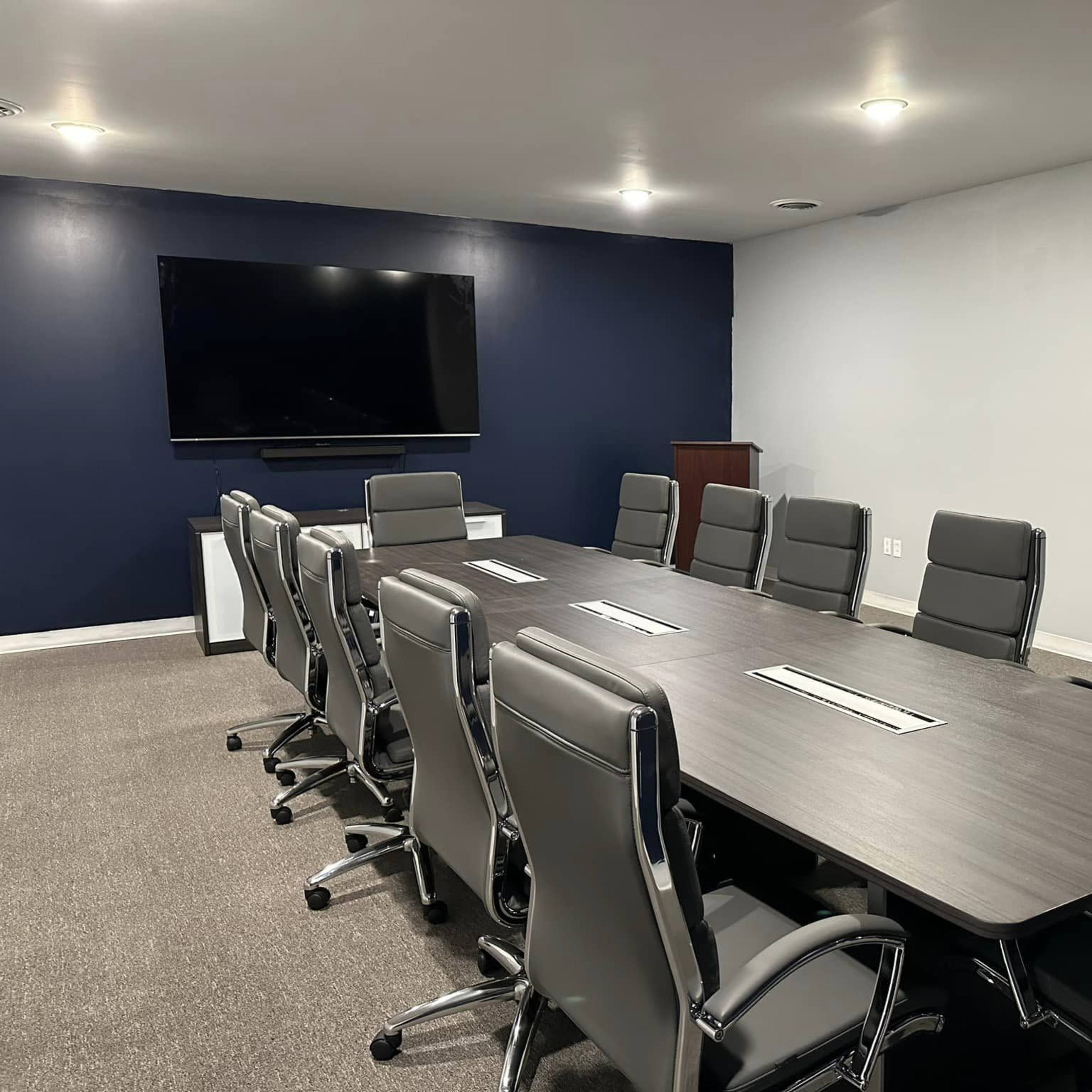 A modern conference room features a long wooden table surrounded by eight ergonomic chairs, with a large screen mounted on the wall.