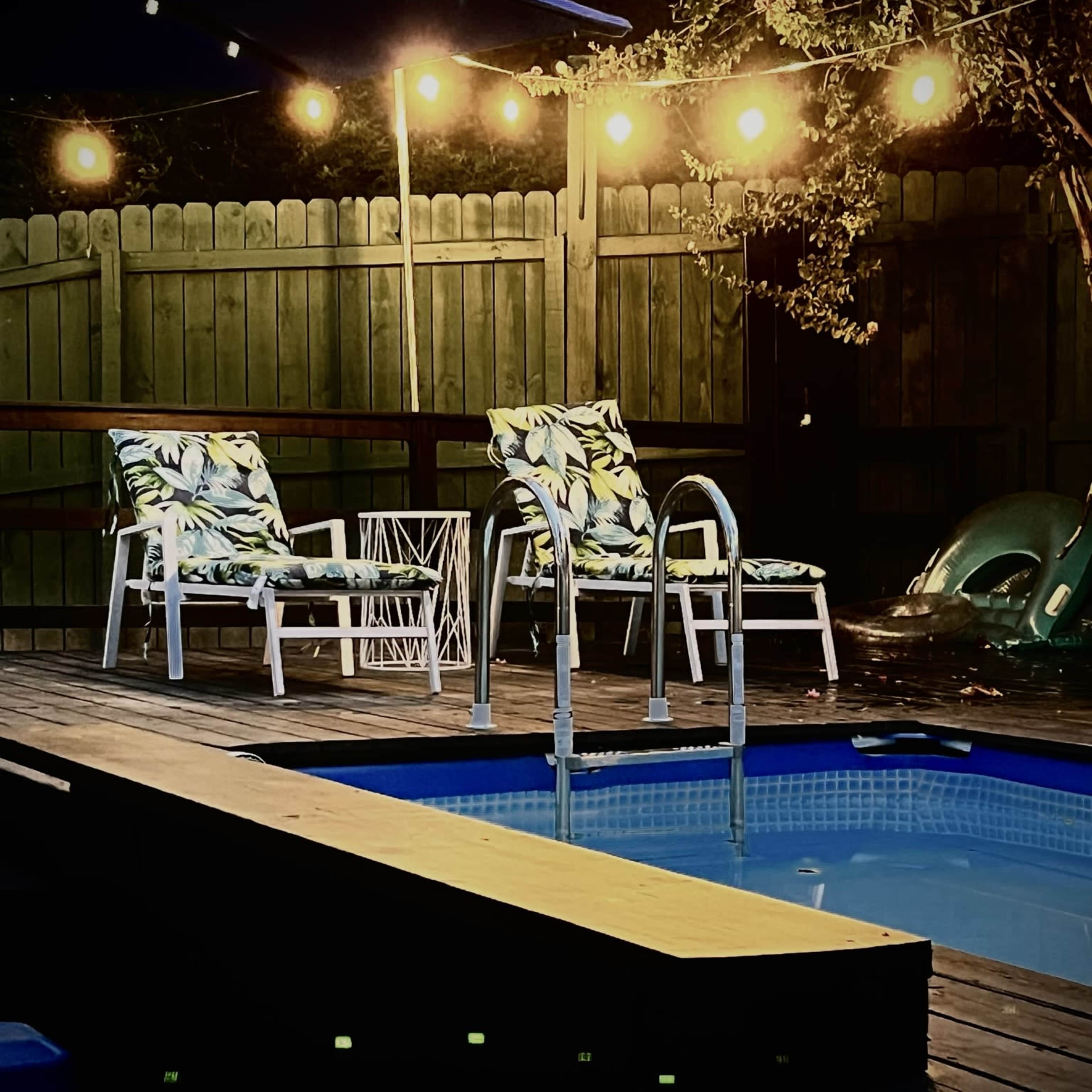 The scene features a swimming pool surrounded by patio chairs, a small table, and string lights illuminating the area at dusk.